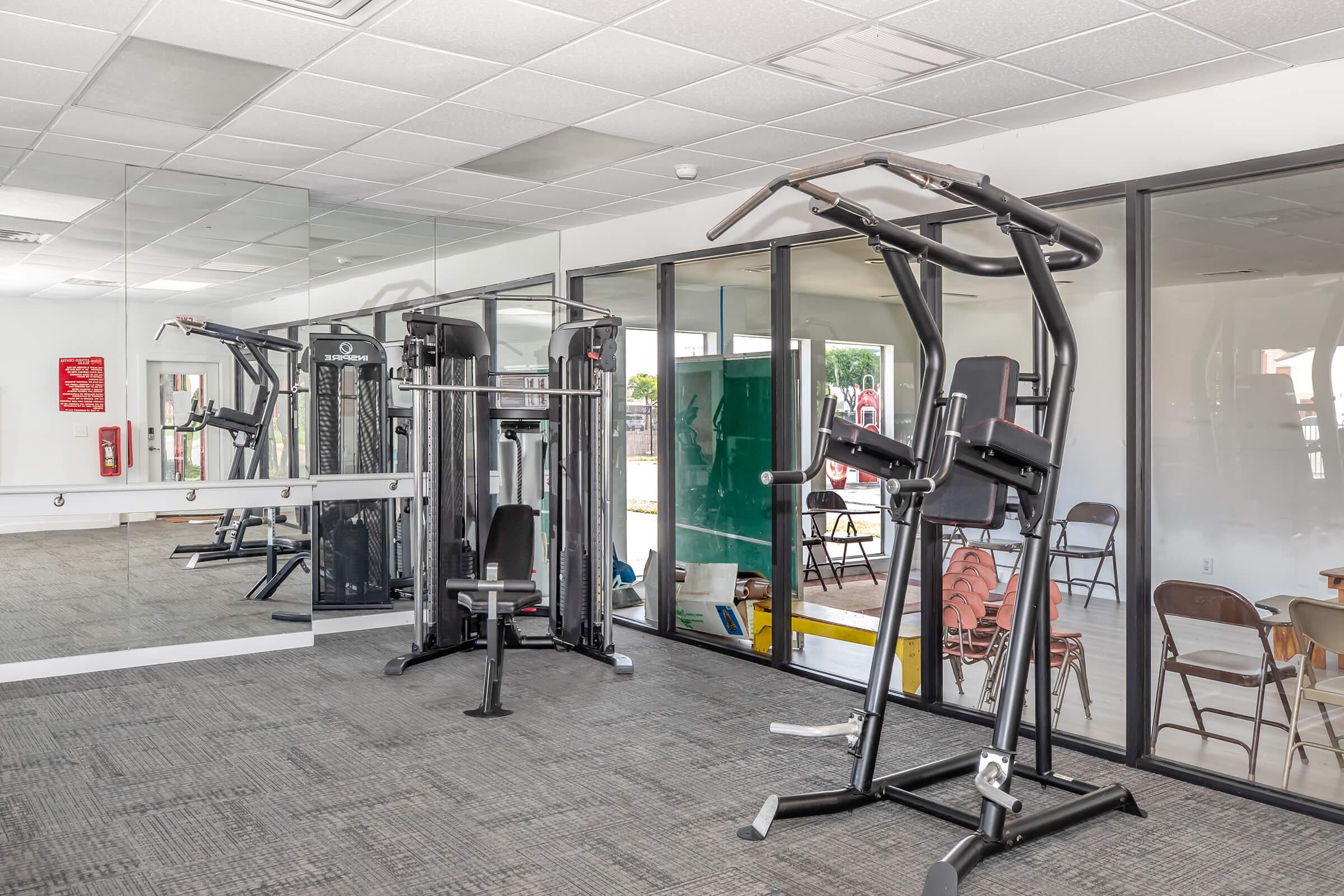 A modern gym interior featuring exercise equipment, including a multi-station weight machine and an elliptical trainer. Large windows provide natural light, and a mirrored wall reflects the space. The flooring is carpeted, and there are empty chairs visible in the background.