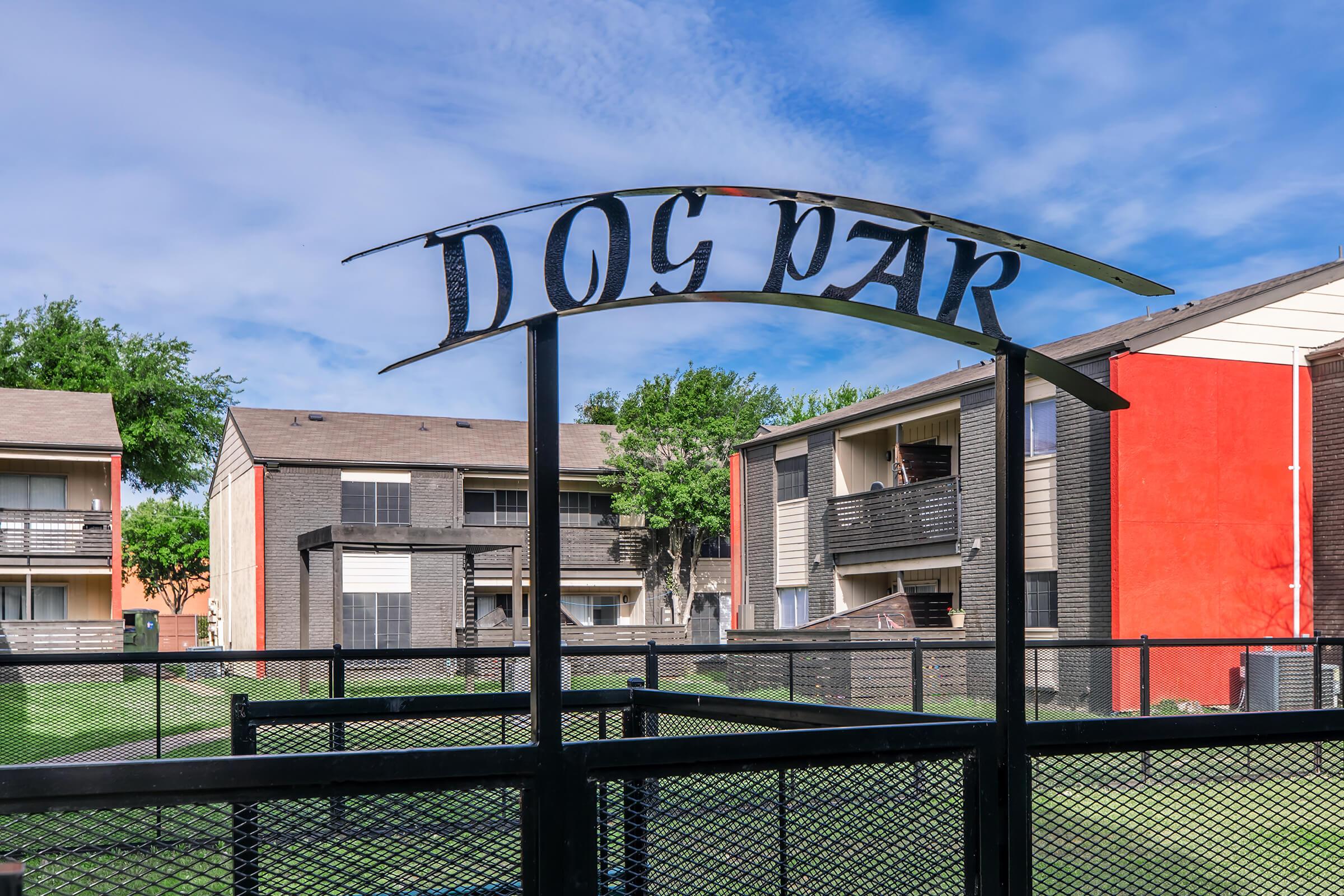 Sign for a dog park with a black metal archway, surrounded by green grass and residential buildings in the background. The sky is partly cloudy, creating a bright and inviting atmosphere.