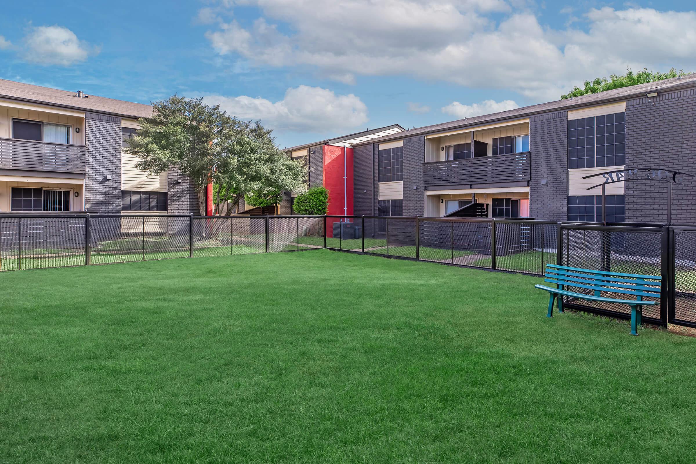 A view of a well-maintained apartment complex featuring two multi-story buildings with dark brick exteriors. In the foreground, there is a green lawn enclosed by a black fence and a blue bench, while trees and shrubs enhance the landscape. Cloudy skies are visible overhead, adding to the serene atmosphere.