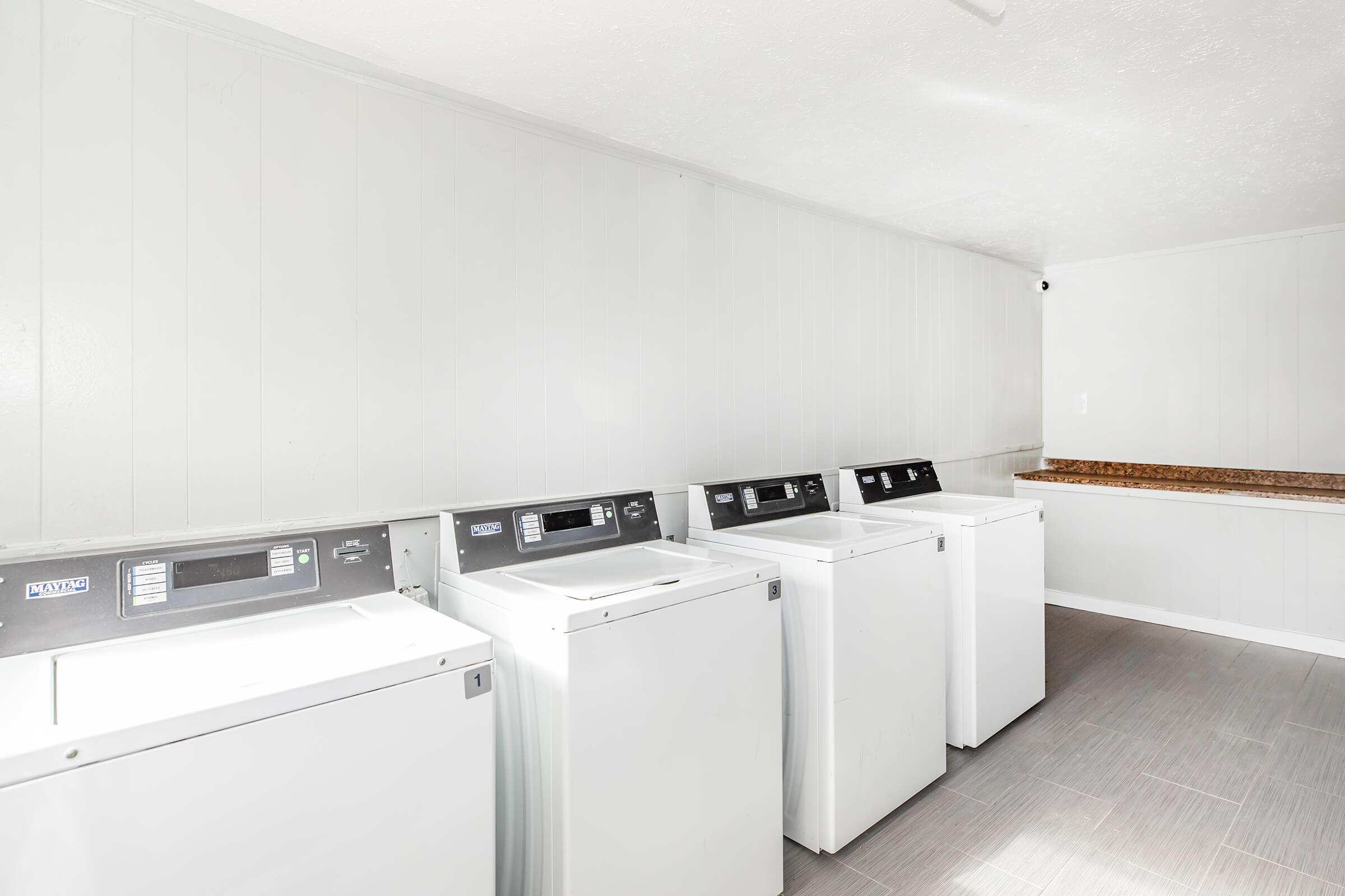 A clean and well-lit laundry room featuring five washing machines lined up against a light-colored wall. There is a countertop along one side, providing space for folding laundry. The floor is tiled in a neutral shade, creating a tidy and functional space.