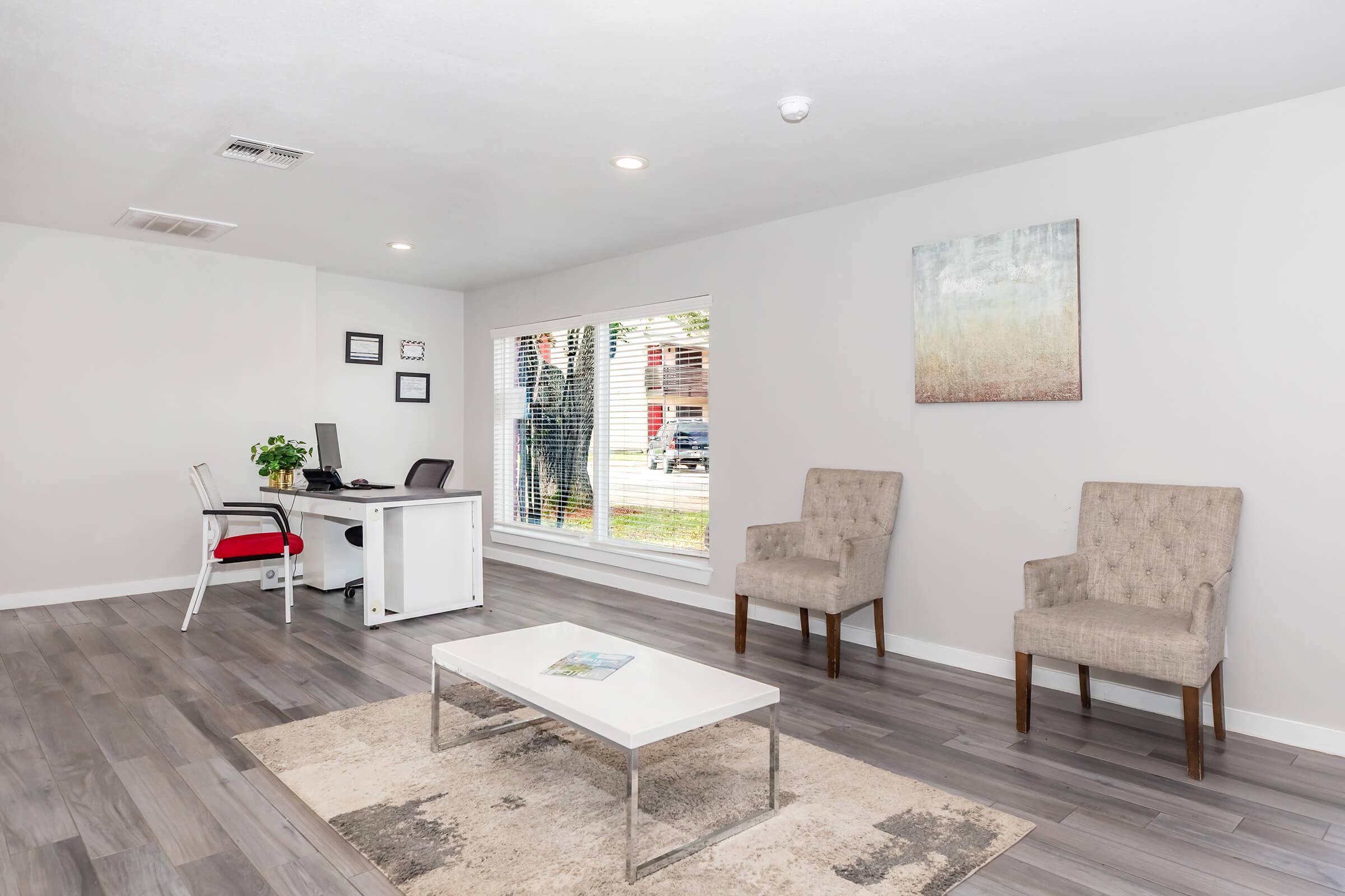 A modern, minimalist office interior featuring a white desk with a chair, two stylish armchairs, and a coffee table on a light-colored rug. There is a large window allowing natural light, and greenery added with a small plant. The walls are painted in neutral tones, creating a calm atmosphere.