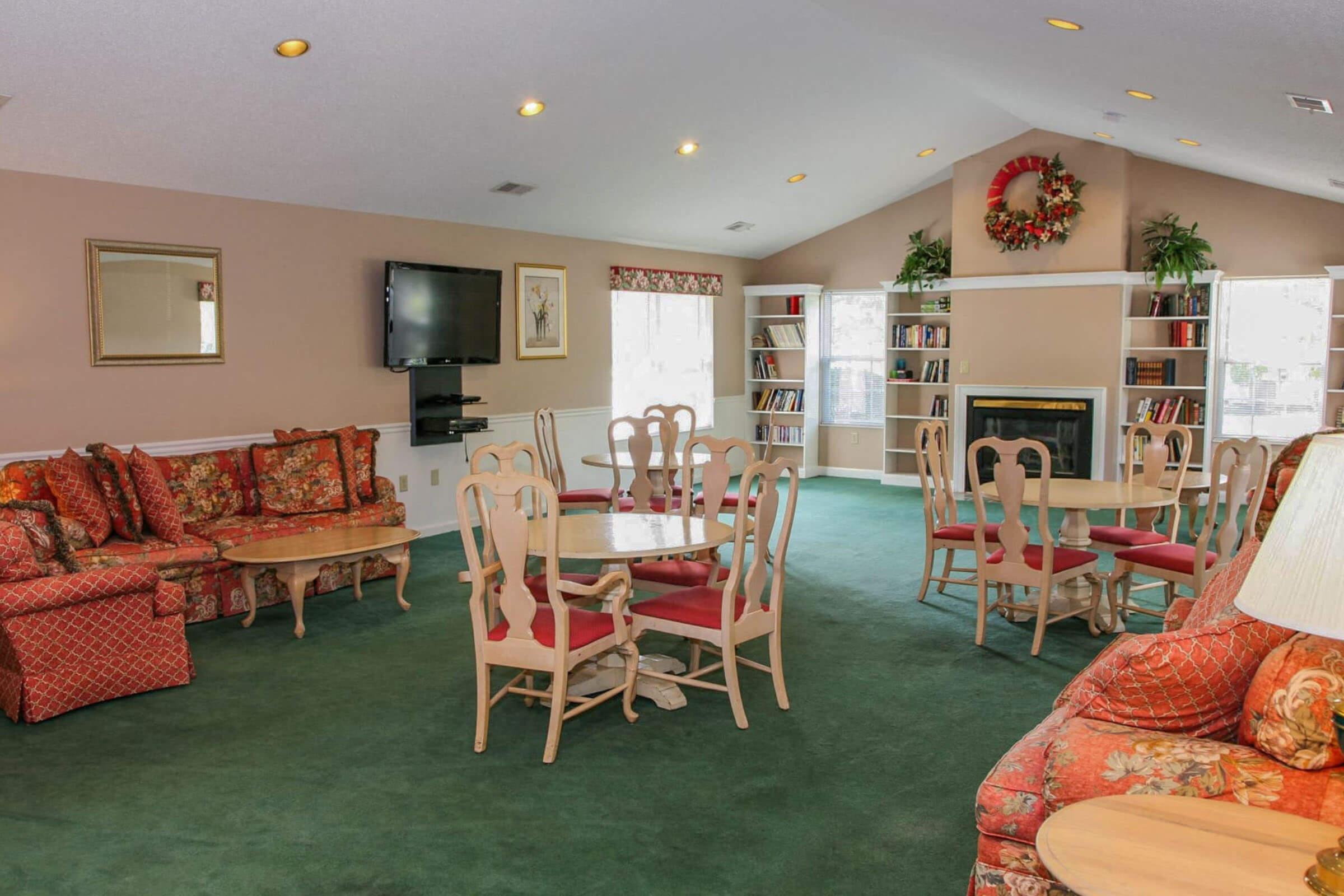 Cozy common area with floral-patterned furniture, including sofas and chairs, arranged around wooden tables. A television is mounted on a wall, and a decorative wreath hangs above a fireplace. Bookshelves filled with books are visible, along with large windows allowing natural light to illuminate the space.