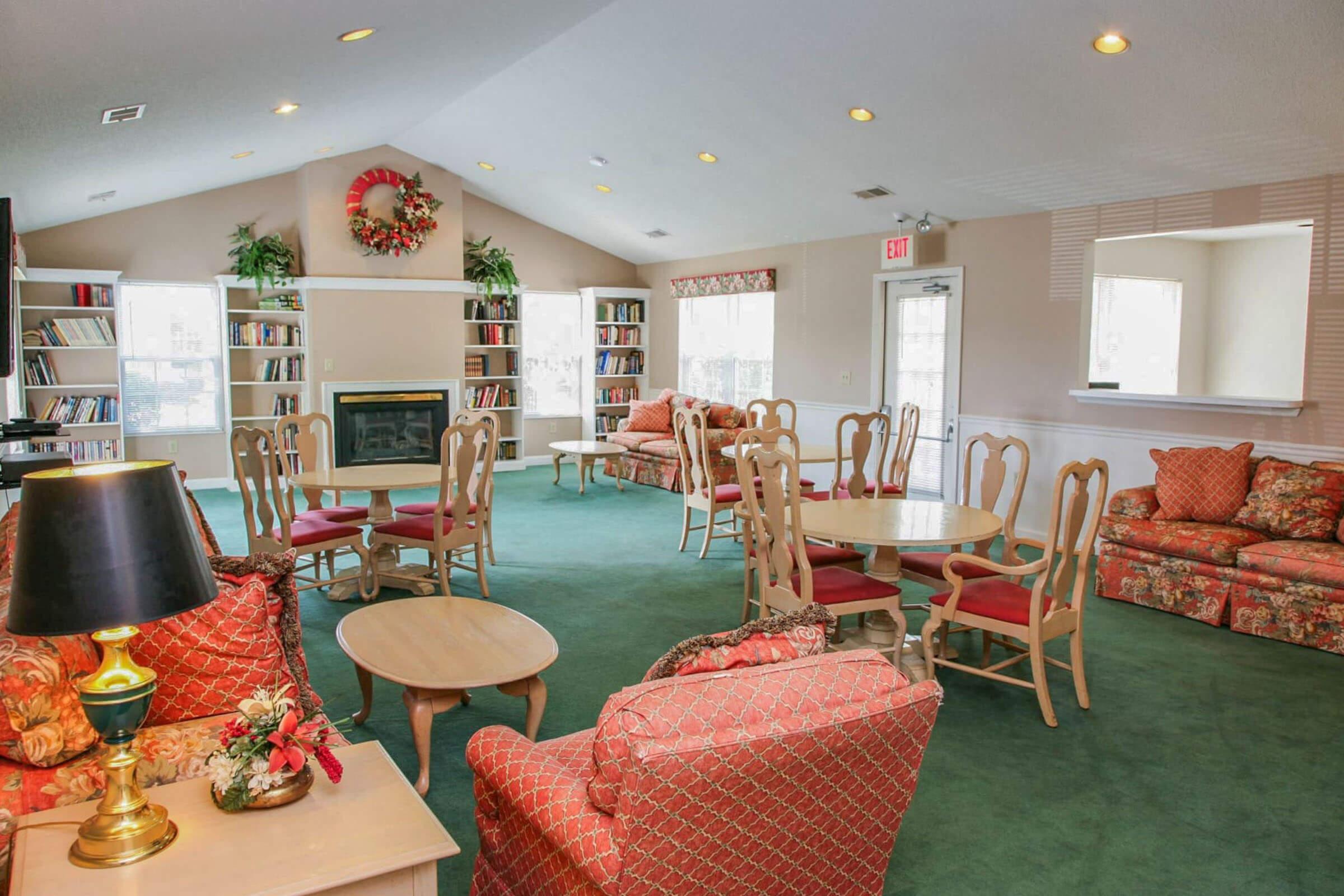 A cozy lounge area featuring comfortable seating, wooden tables, and bookshelves. The decor includes vibrant floral patterns and a festive wreath on the wall. Soft lighting and large windows enhance the welcoming atmosphere, making it an inviting space for relaxation or reading.