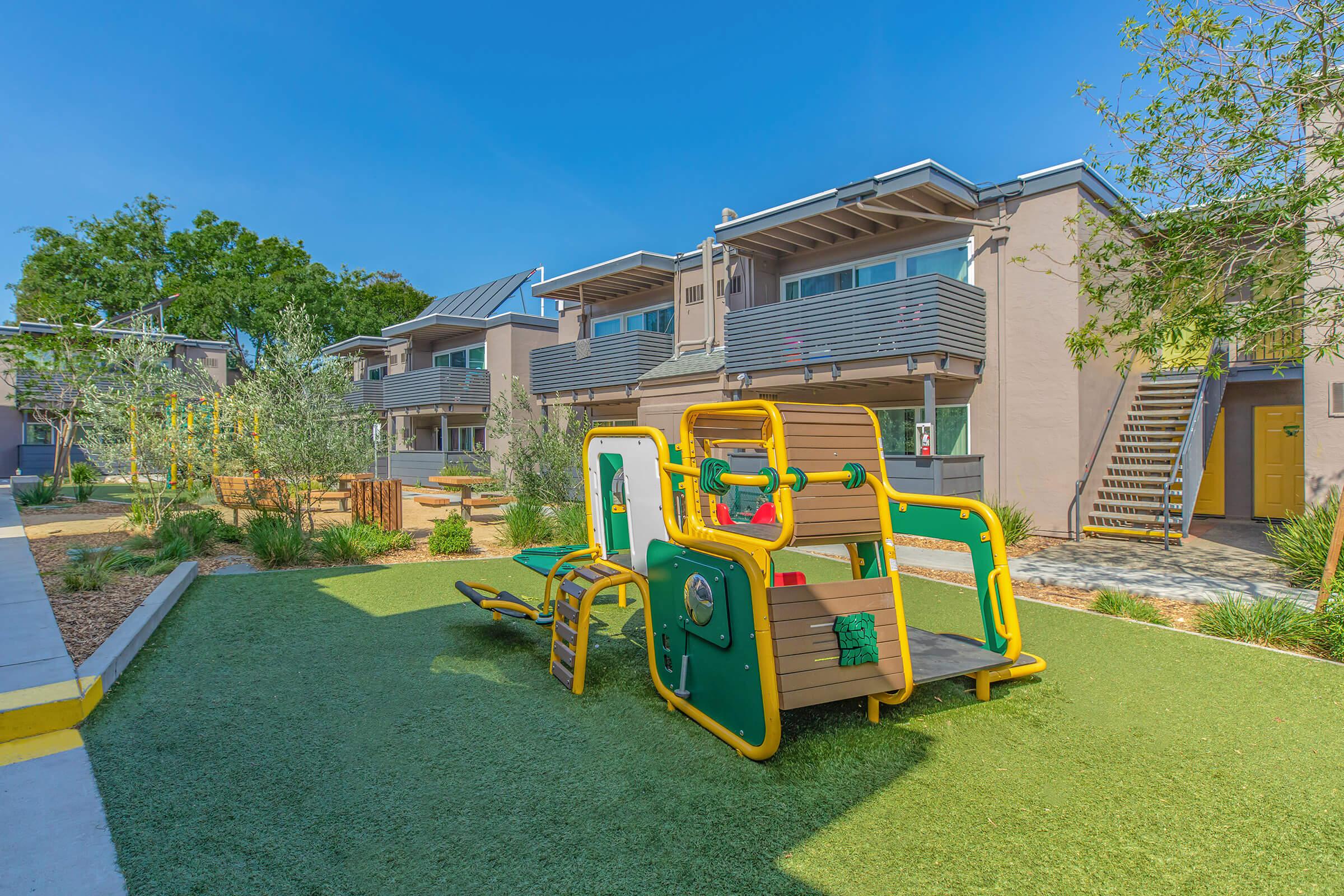 Play area equipment with green outdoor flooring and landscaping, background of apartment buildings.