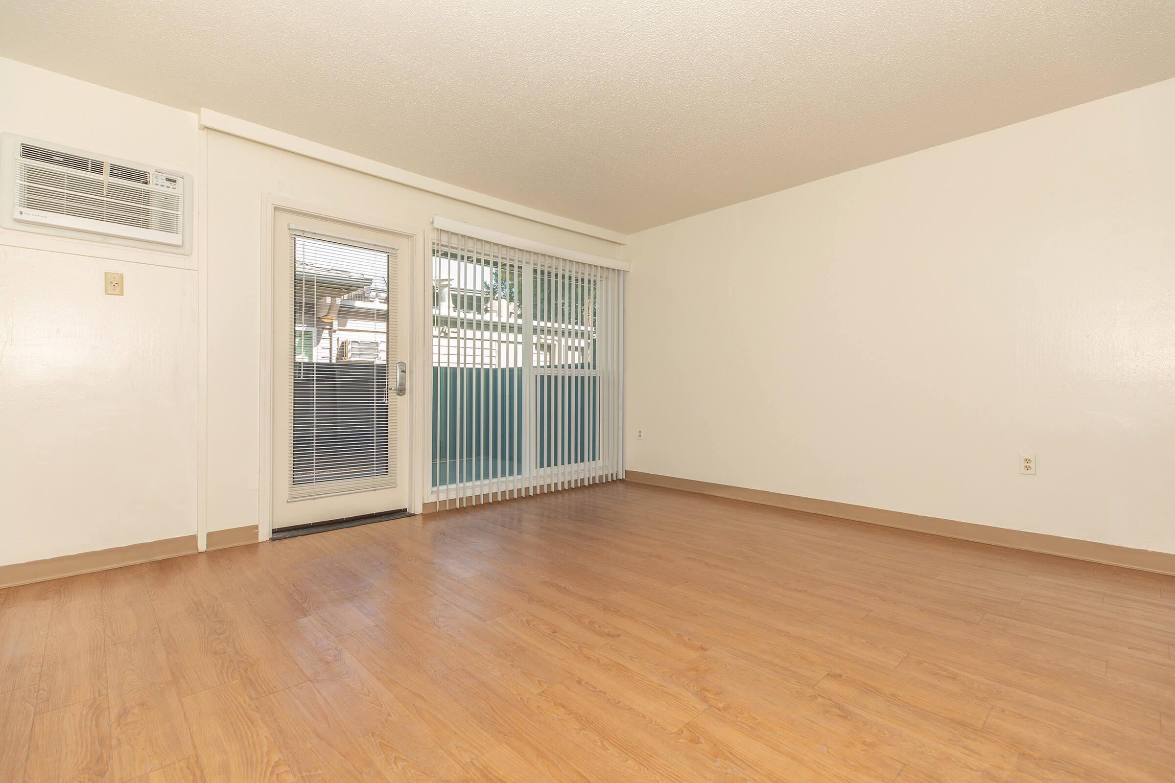 Apartment living room with wall air conditioning unit and door leading outside next to floor-to-ceiling windows.