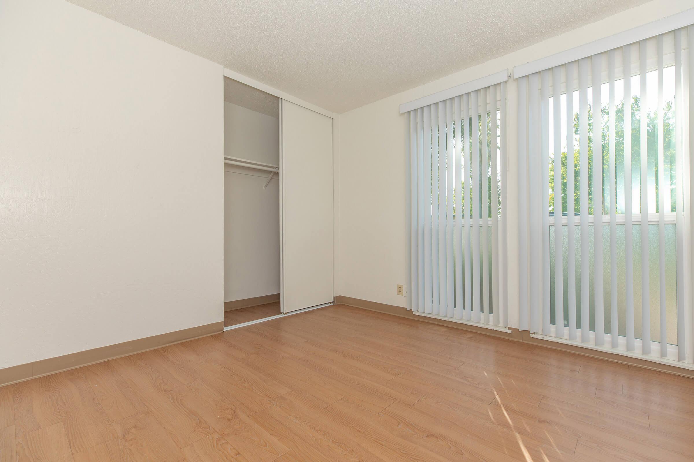 Bedroom with open closet, tall windows with vertical blinds, white walls, and hardwood floors.