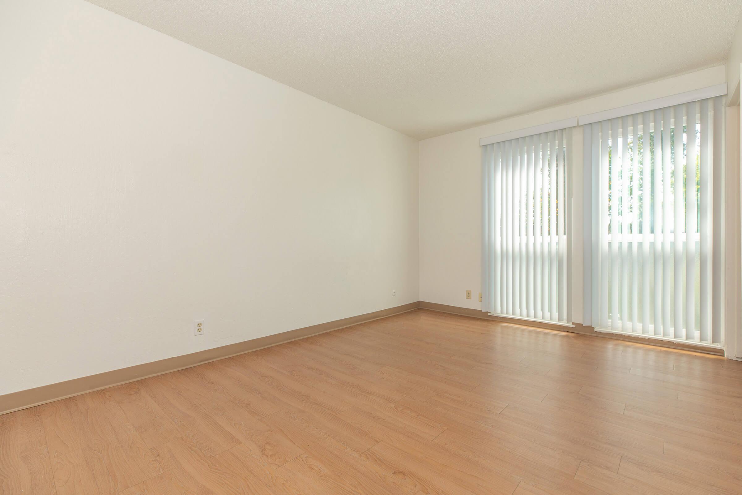 Bedroom with tall windows, white walls, hardwood floors, and vertical blinds.