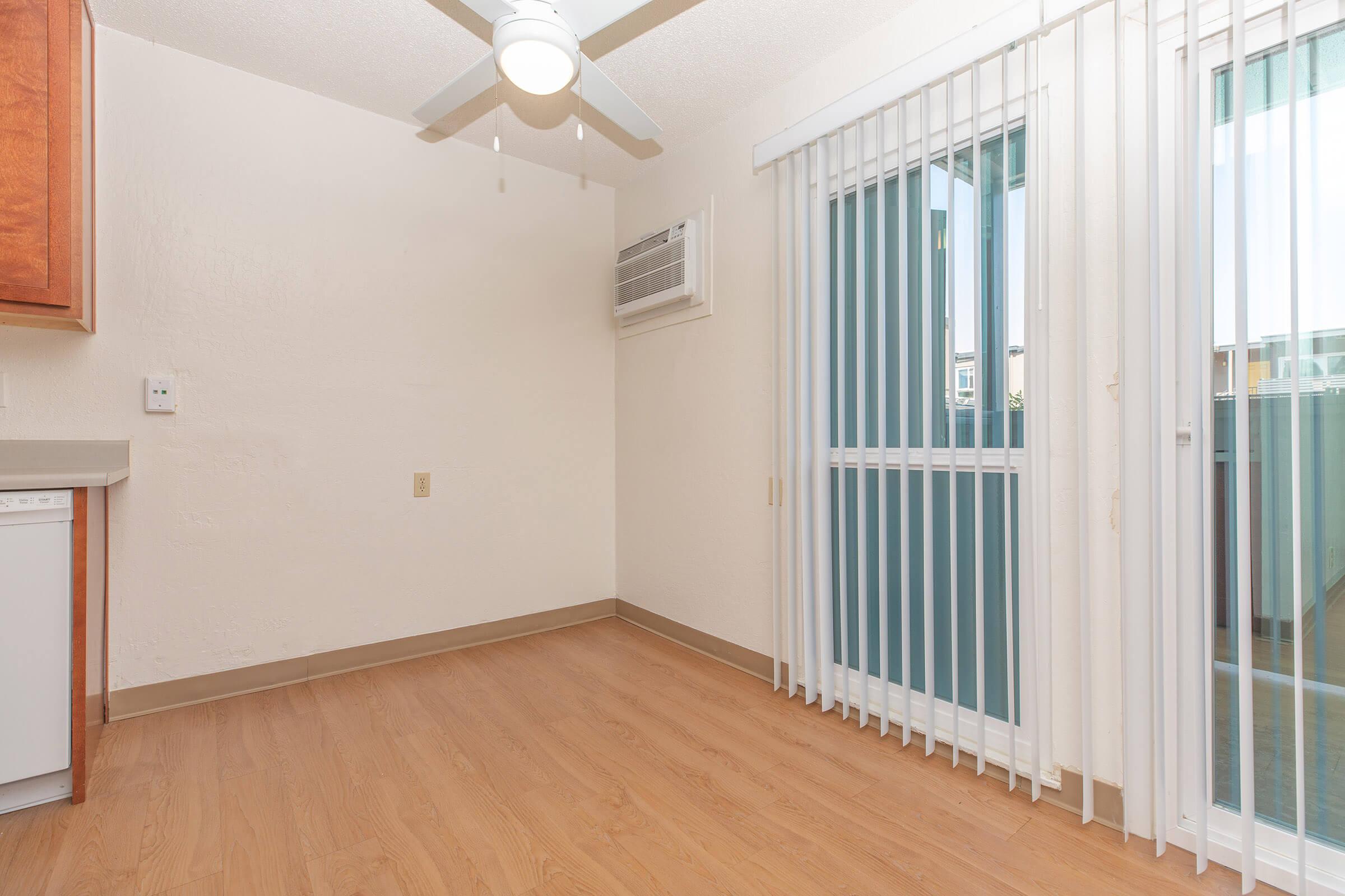 Dining area with white walls, hardwood floors, ceiling fan, wall air conditioning unit, and tall windows with vertical blinds.