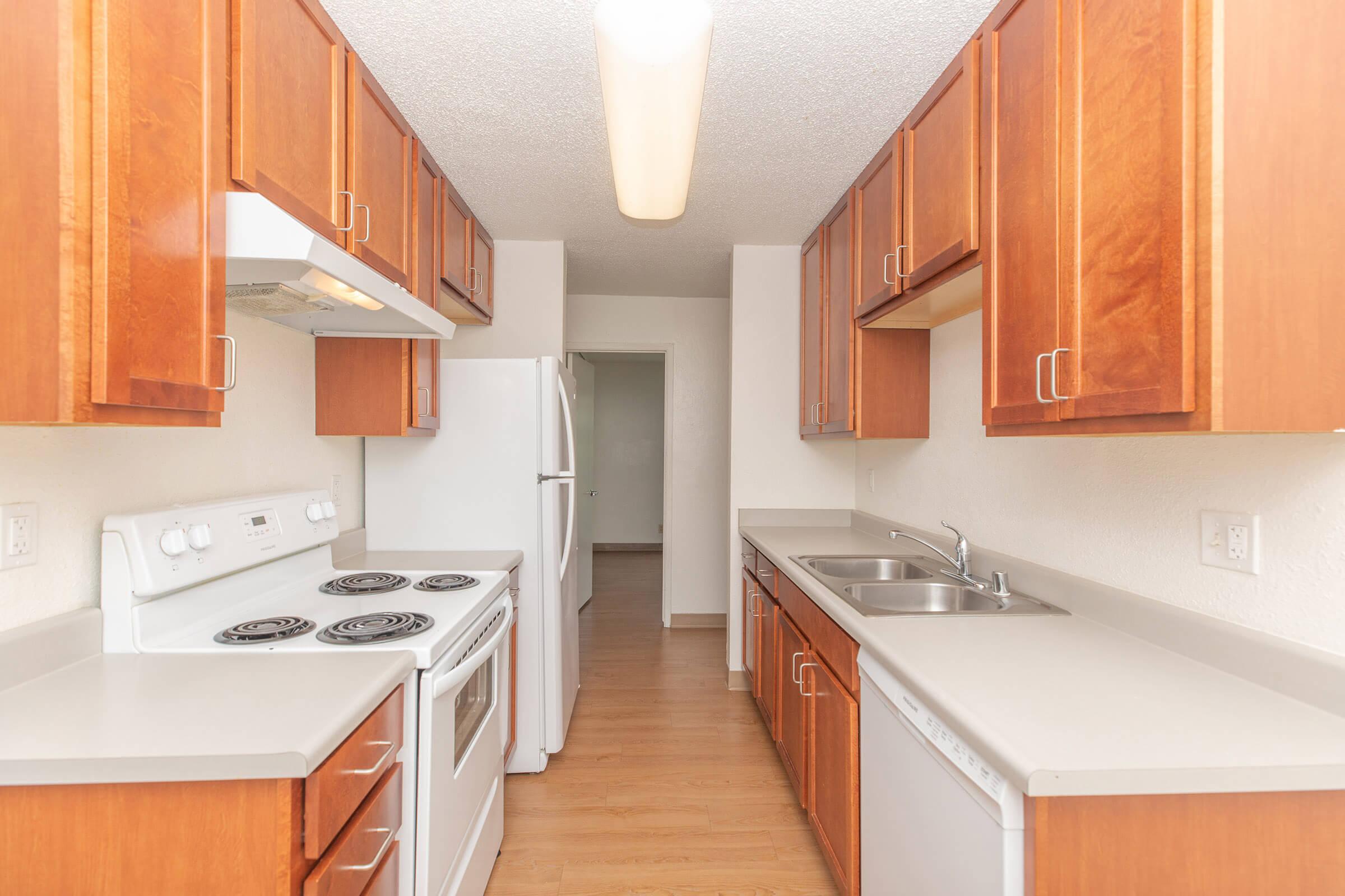 Two bedroom apartment galley kitchen with white appliances and cherry colored wood cabinetry, view into small room.