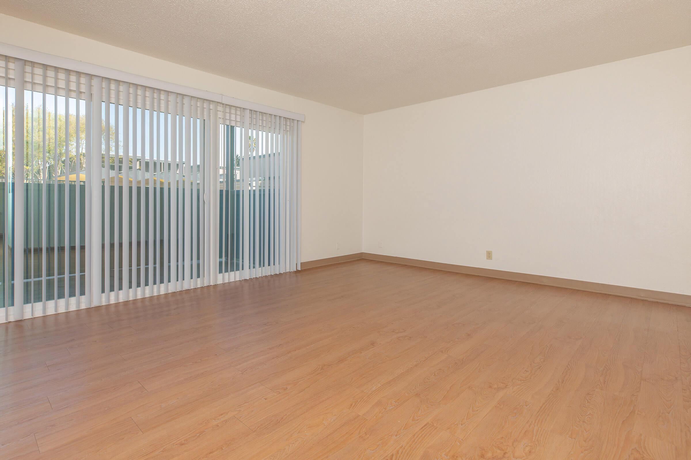 Two bedroom apartment living room with white walls, hardwood floors, and tall windows with vertical blinds.