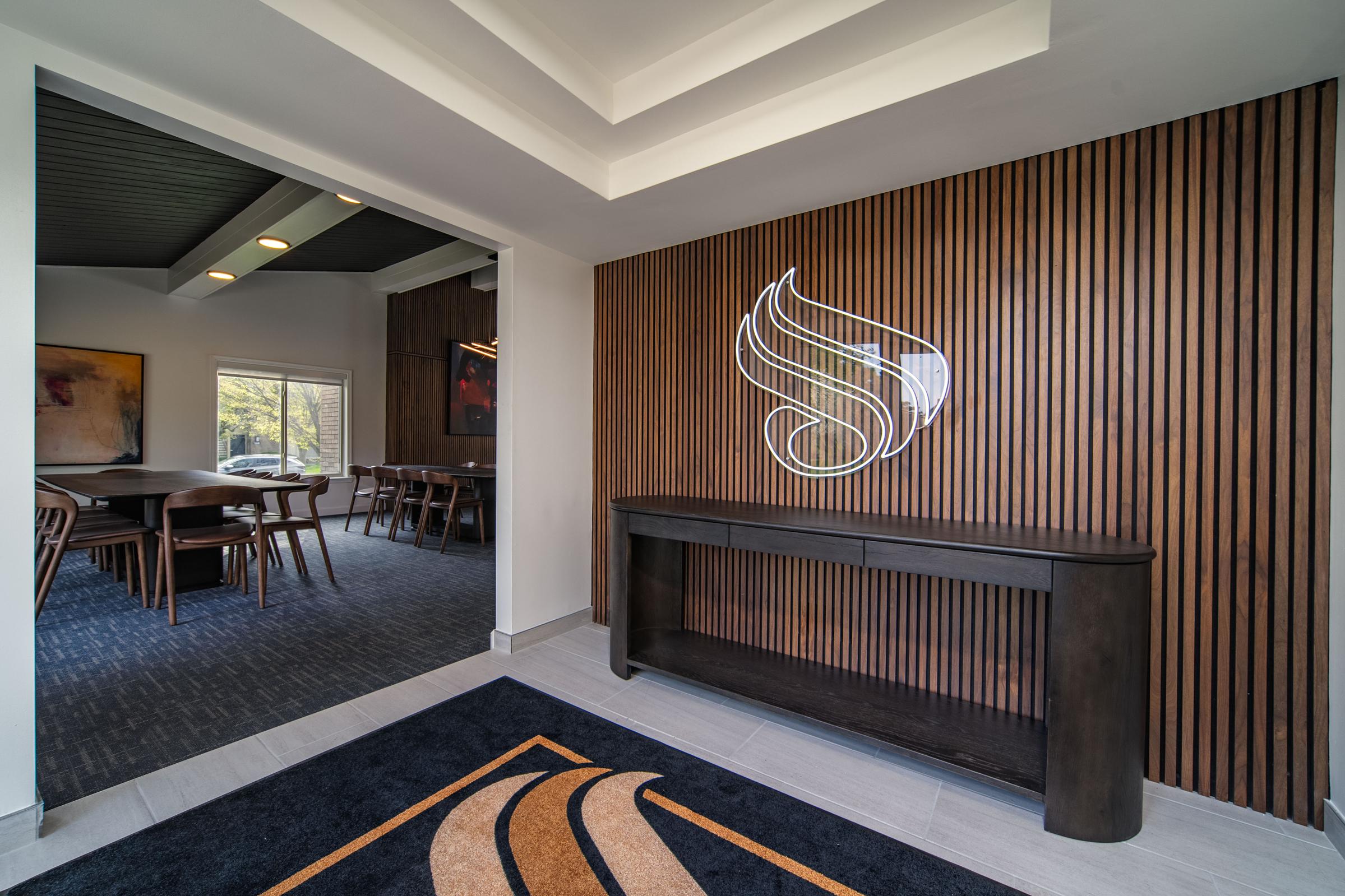 Interior view of a modern lobby featuring a wooden accent wall, a sleek dark console table, and a stylized logo. Natural light filters through a nearby window, illuminating a dining area with chairs and tables in the background. The overall decor combines contemporary and cozy elements.