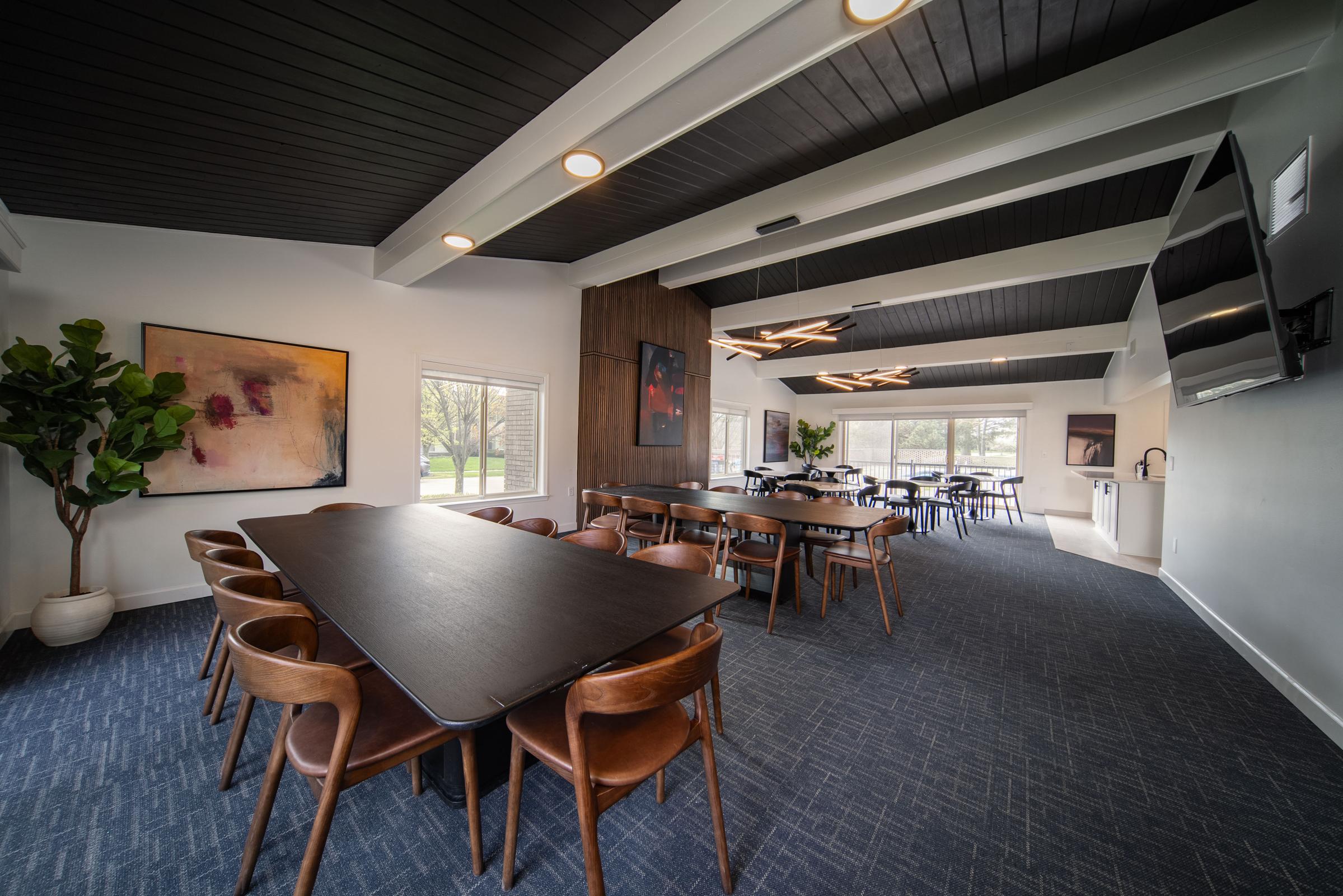 A modern meeting room featuring a long black table surrounded by wooden chairs. Large windows allow natural light to fill the space, which includes contemporary art on the walls and a green plant in a pot. There’s also a television and a kitchenette area, providing a comfortable setting for gatherings.