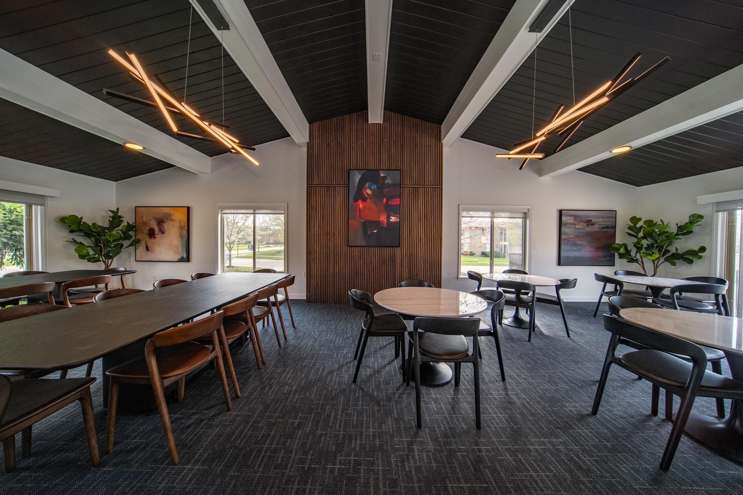 Interior of a modern meeting room featuring a large wooden table and several smaller round tables. The space has a high ceiling with stylish light fixtures, large windows, and potted plants. Abstract artwork adorns the walls, creating a contemporary and inviting atmosphere.