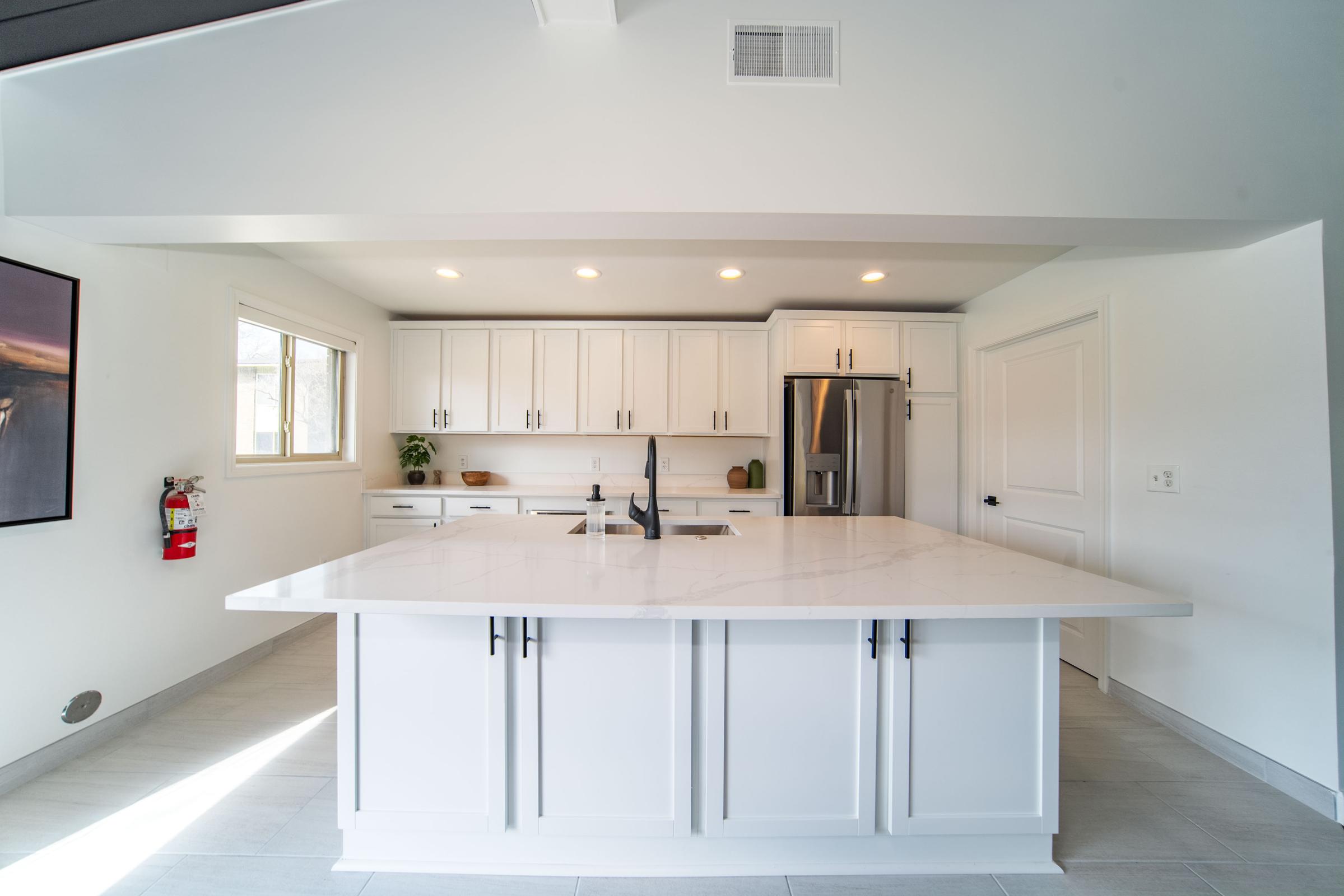 A modern kitchen featuring white cabinetry, a large marble island with a sink, stainless steel appliances, and natural light coming from a window. The space has a clean and minimalist design with gray tile flooring. A fire extinguisher is mounted on the wall for safety.