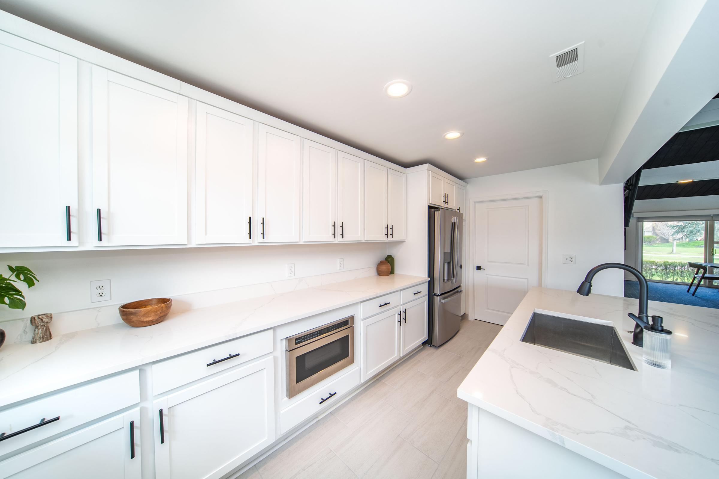 Modern kitchen featuring white cabinetry with black hardware, a spacious countertop with a sink, built-in microwave, and stainless steel refrigerator. The design is bright and open, with natural light streaming in from a nearby window. A clean and minimalistic aesthetic creates a fresh ambiance.