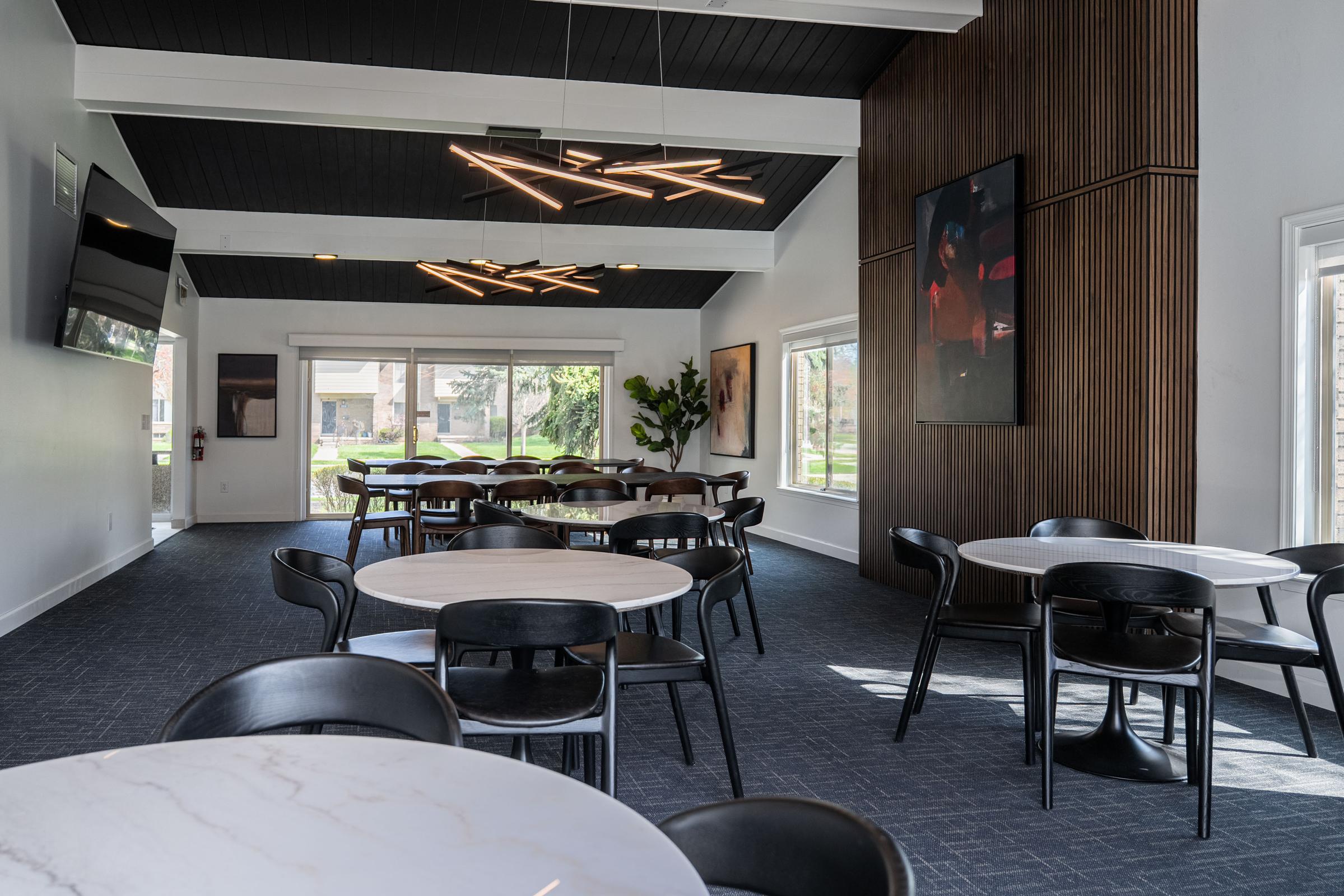 Interior view of a modern dining or meeting space featuring several round tables with black chairs, sleek lighting fixtures, and large windows. Decorative elements include a wooden accent wall and artwork. The room has a contemporary feel with a neutral color palette and ample natural light.