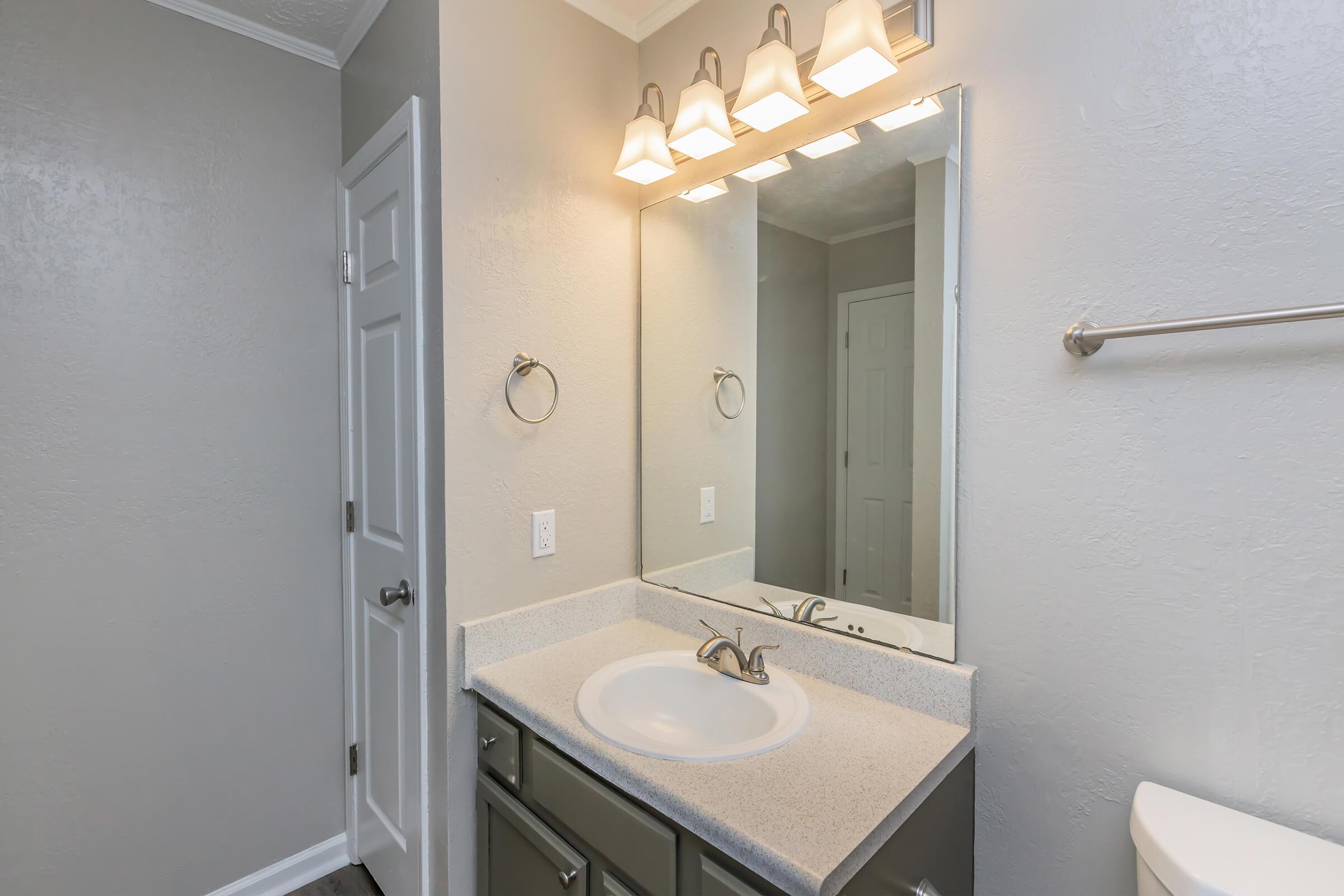A well-lit bathroom featuring a modern vanity with a white sink, gray cabinet, and a large mirror above. The walls are painted in a light gray, and there are five light fixtures above the mirror. A towel ring is mounted on the wall, and a white toilet is visible in the corner.
