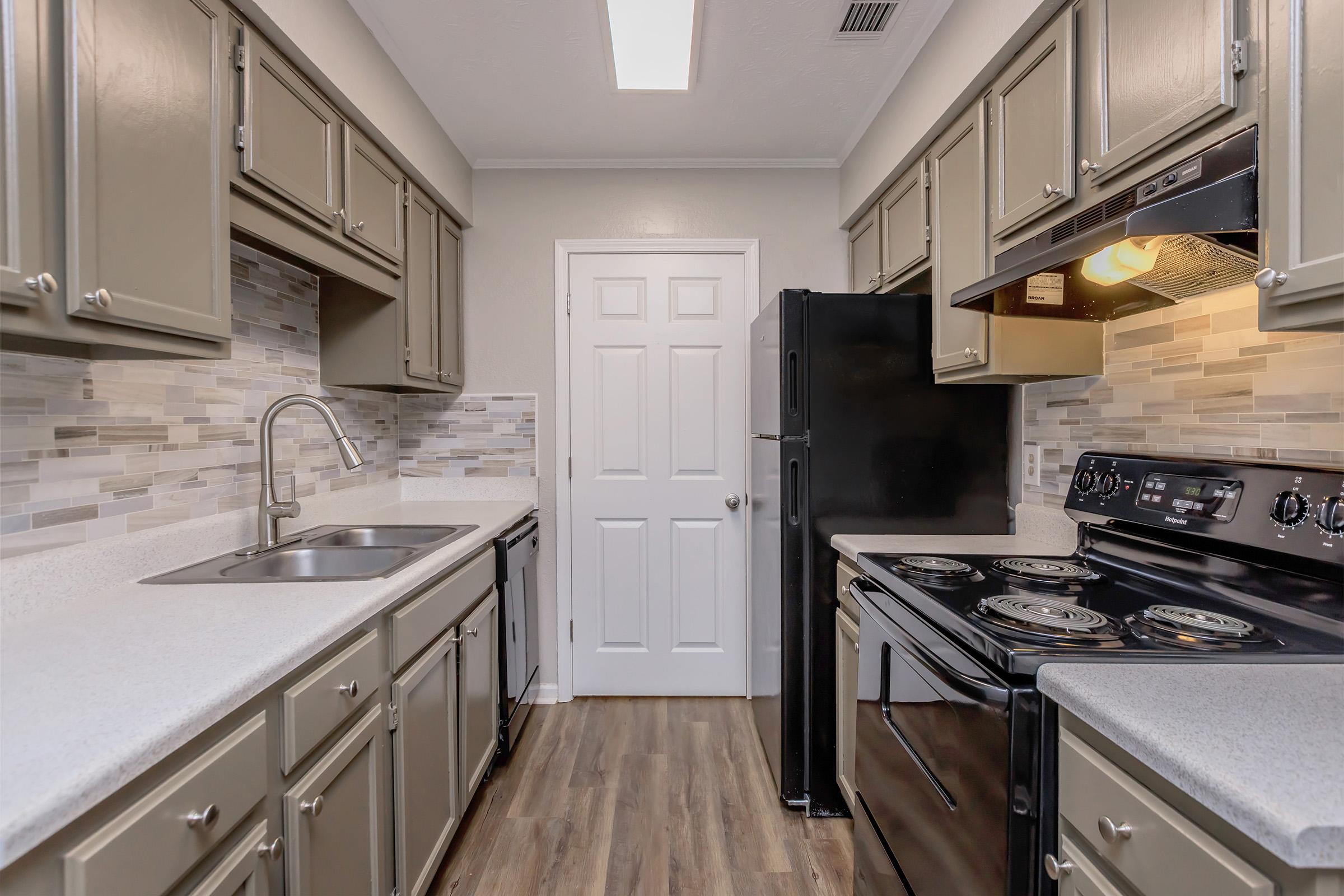 A modern kitchen featuring gray cabinets, a black stove and refrigerator, a stainless steel sink, and a light countertop. The backsplash consists of decorative tiles, and the room has a door leading to another space. Natural light floods in from a ceiling fixture.