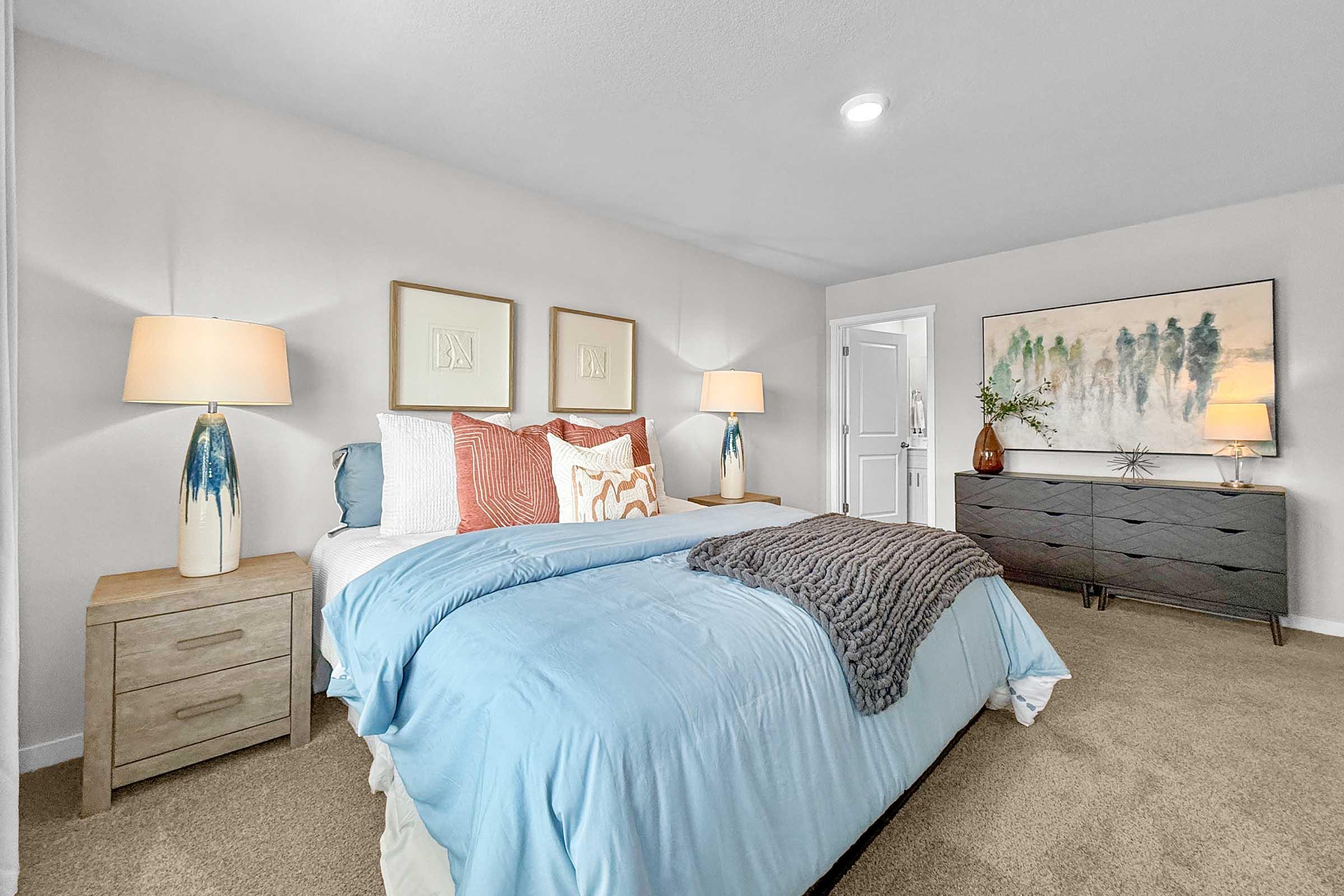 A cozy bedroom featuring a light blue bedspread with decorative pillows, two bedside lamps on wooden tables, and a dark dresser. The walls are painted in a soft white, with framed artwork and a vibrant landscape painting above the dresser, creating a serene and inviting atmosphere.