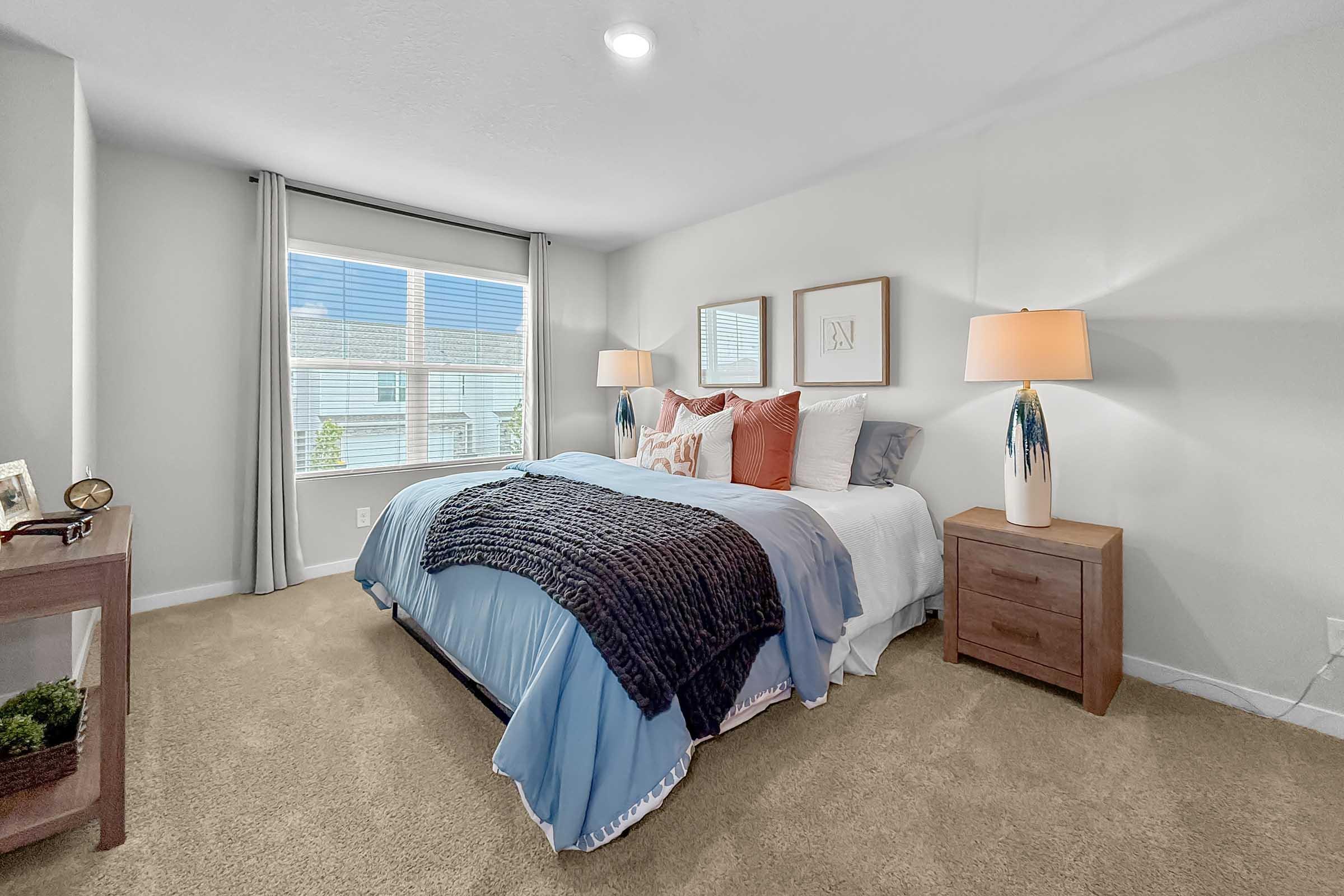 Cozy bedroom featuring a queen-sized bed with light blue and white bedding, accented by decorative pillows. Two bedside lamps provide warm lighting, and a window allows natural light to brighten the space. A small wooden nightstand and a decorative piece add charm to the room. Light beige carpet completes the inviting atmosphere.