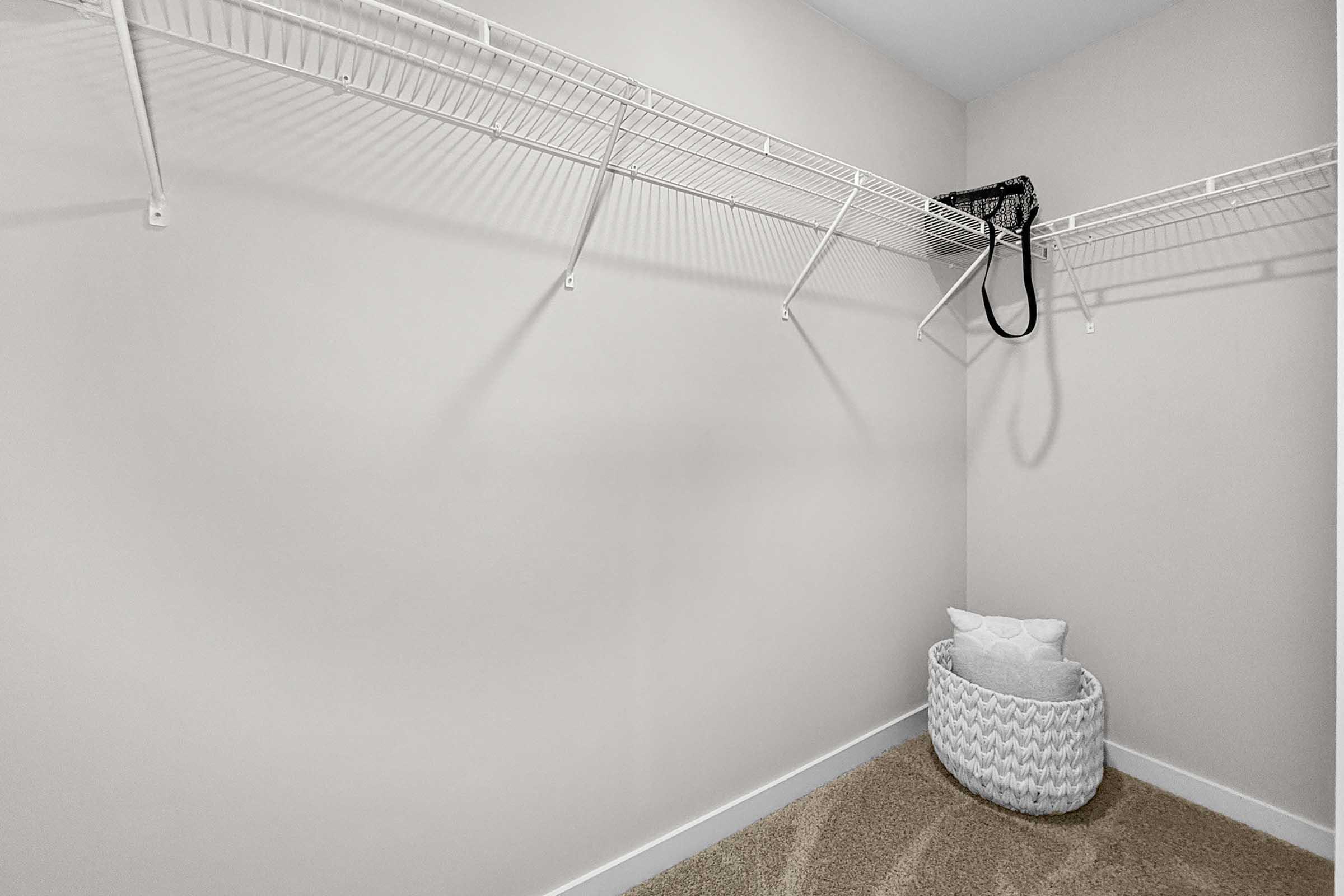A minimalist closet with empty wire shelving against a plain wall. A black bag hangs from the shelf, and a woven basket sits on the floor, next to a textured carpet. The space is well-lit and organized, conveying a clean and tidy appearance.