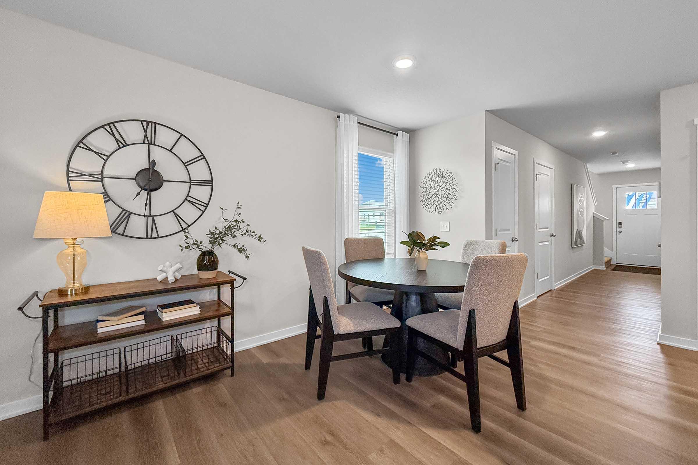 A modern dining area featuring a round table with four upholstered chairs. A stylish wall clock is mounted on a neutral-colored wall, alongside a small console table with decor. Soft lighting from a lamp creates a cozy atmosphere, and a doorway leads to an entrance and staircase in the background.