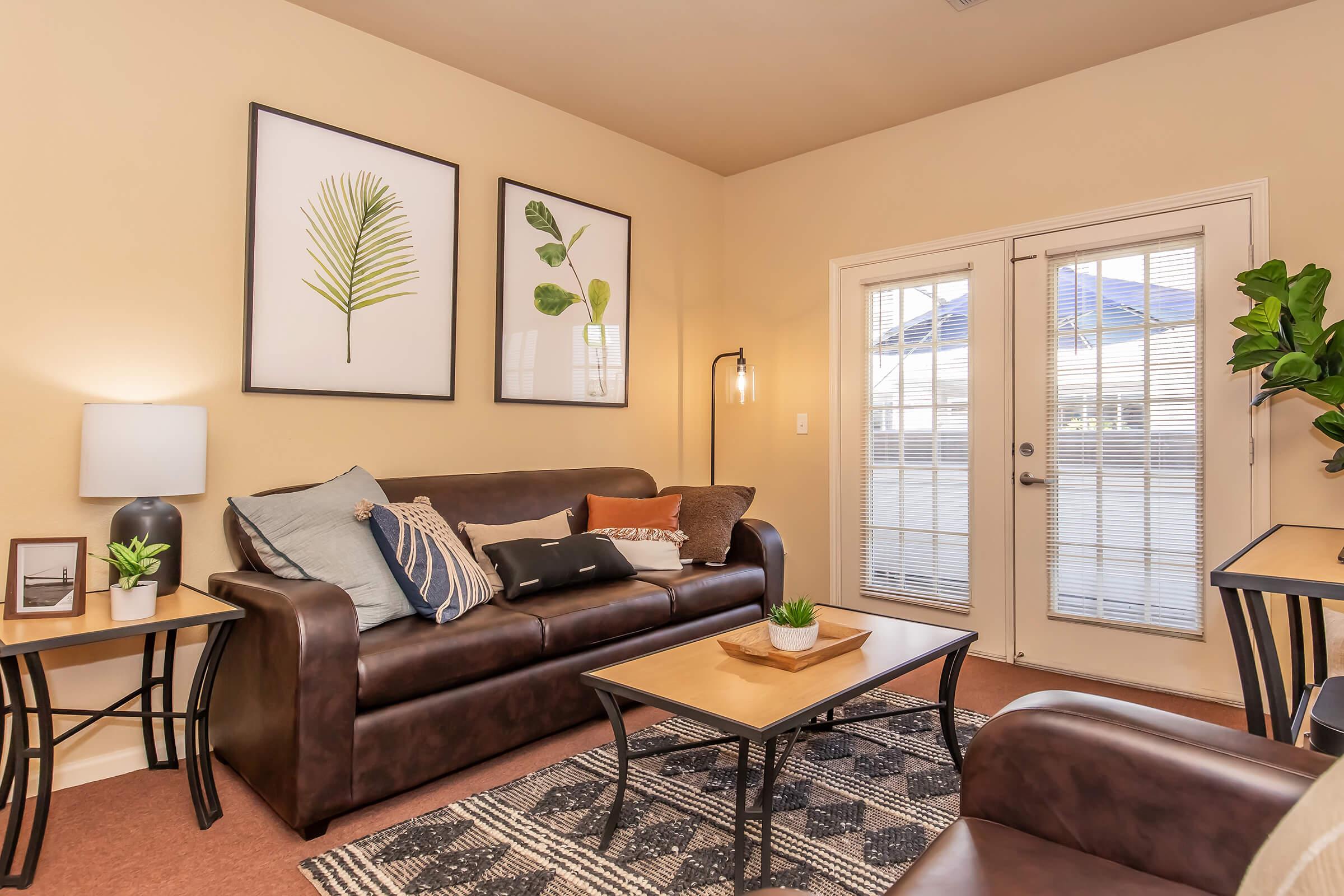 A cozy living room featuring a brown leather sofa with decorative pillows, a wooden coffee table, and a side table with a lamp. The walls are adorned with framed botanical prints, and a large window with blinds allows natural light to enter. A potted plant adds a touch of greenery to the space.
