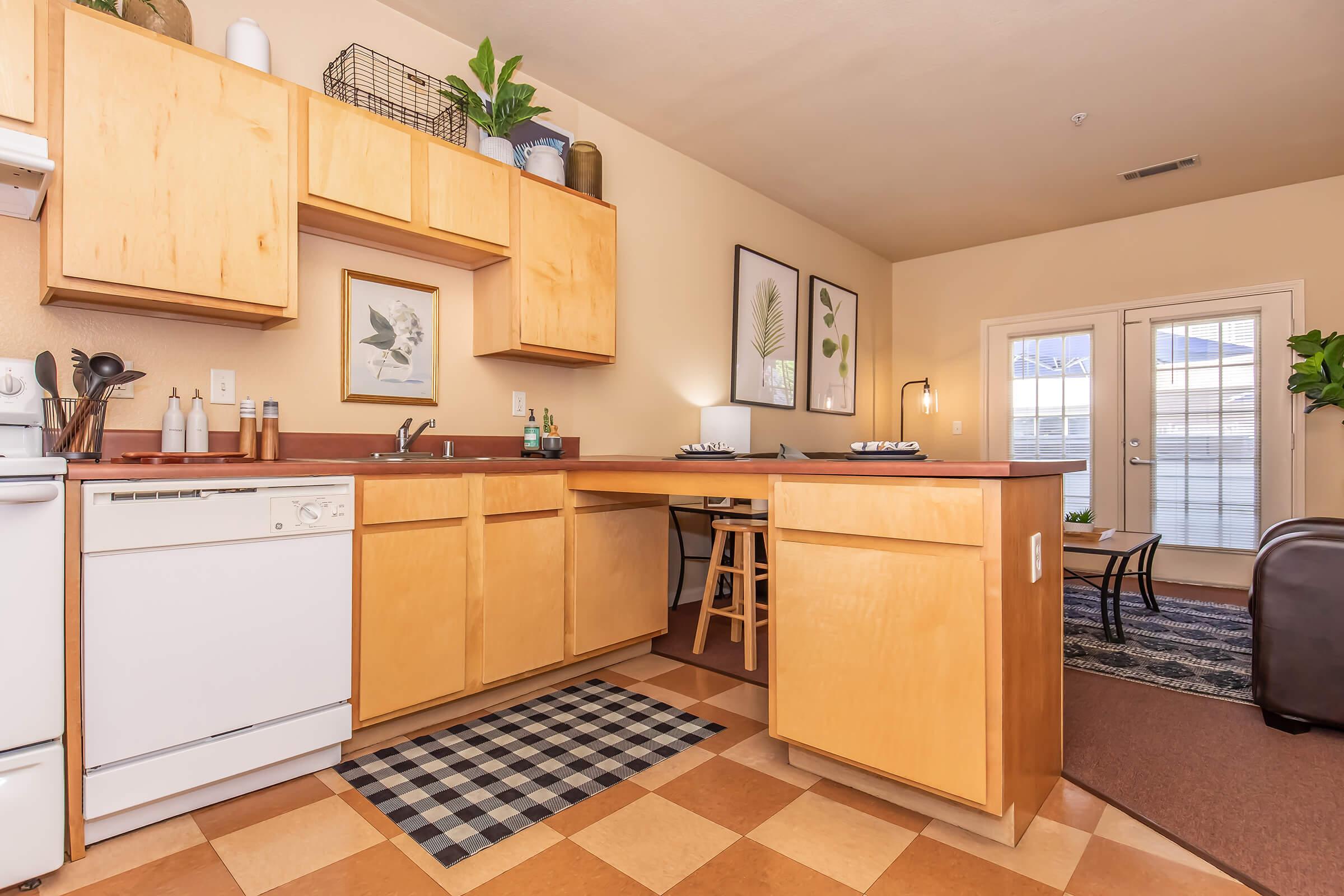 A cozy kitchen featuring light wood cabinetry, a white dishwasher, and a sink. The space includes a small dining area with a stool, a decorative black and white checkered rug, and framed botanical art on the walls. Large windows provide natural light, enhancing the warm, inviting atmosphere of the room.