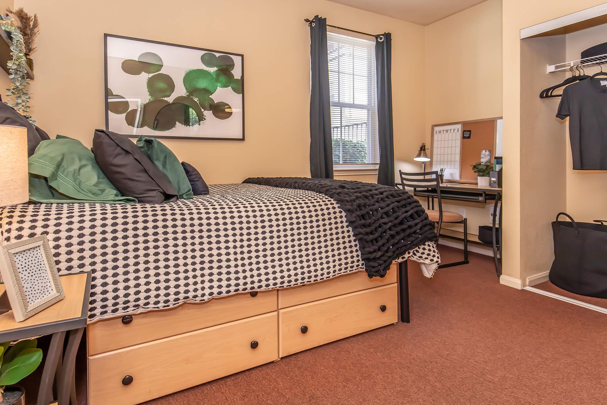 A cozy bedroom featuring a large bed with a patterned black and white comforter, decorative pillows, and a framed art piece on the wall. A small desk with a chair is positioned near a window, alongside a closet with clothing and a mirror. The space is warmly illuminated and has a welcoming atmosphere.