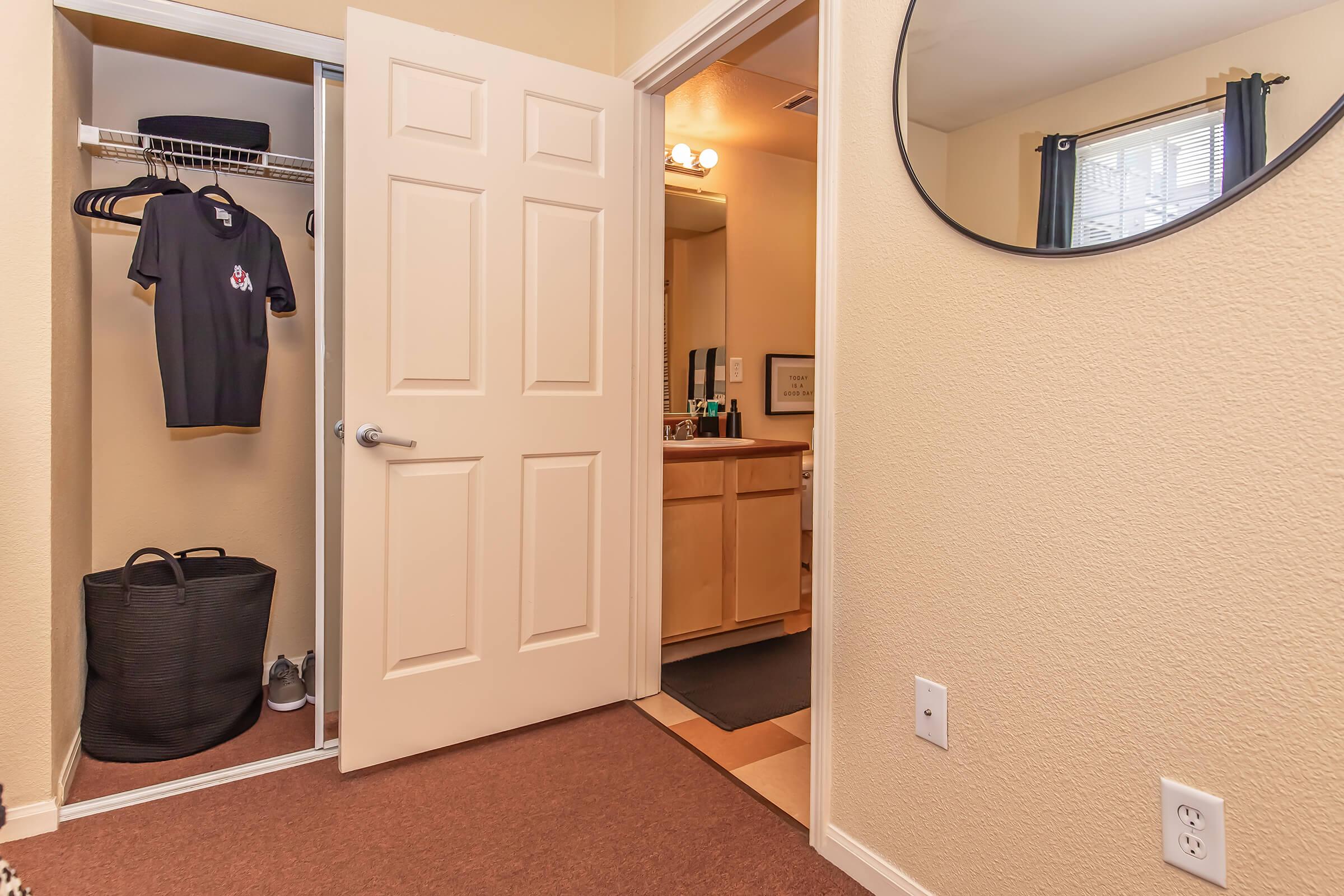 A view of an entryway with a closet on the left featuring a shirt, a laundry basket, and shoes. To the right, a bathroom vanity is partially visible through an open door, with a round mirror above the sink. The walls are painted a light color, and the flooring includes carpet and tile.