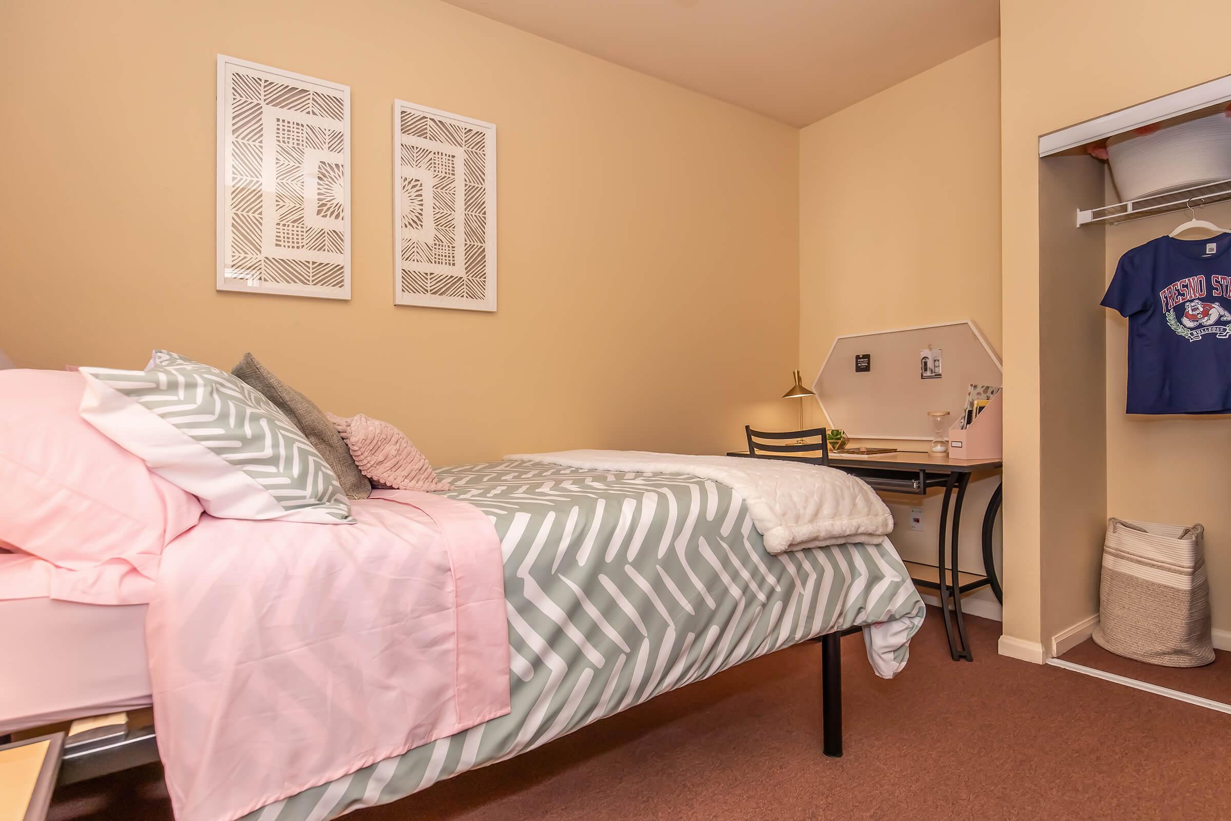 A cozy bedroom featuring a twin bed with a patterned comforter, decorative pillows, and wall art. A small desk with a lamp is situated near the bed, and a closet is visible with a shirt hanging inside. The room has warm beige walls and carpet flooring, creating an inviting atmosphere.