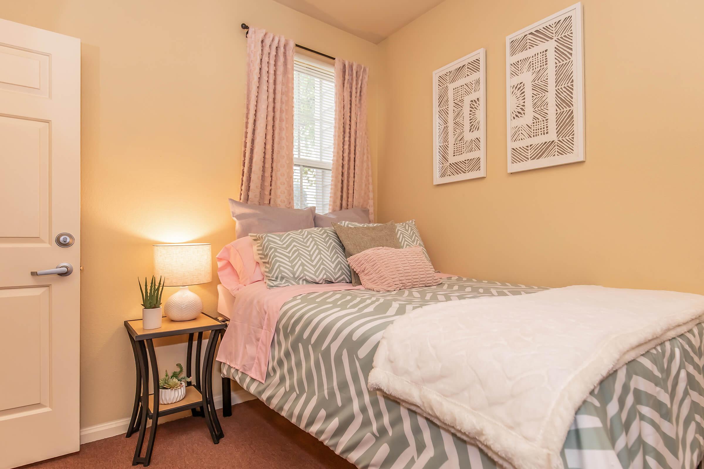 Cozy bedroom featuring a twin bed with striped bedding and decorative pillows. A small bedside table holds a lamp and a potted plant. Light-colored walls and window curtains enhance the warm ambiance, while framed geometric art adds a stylish touch to the decor.