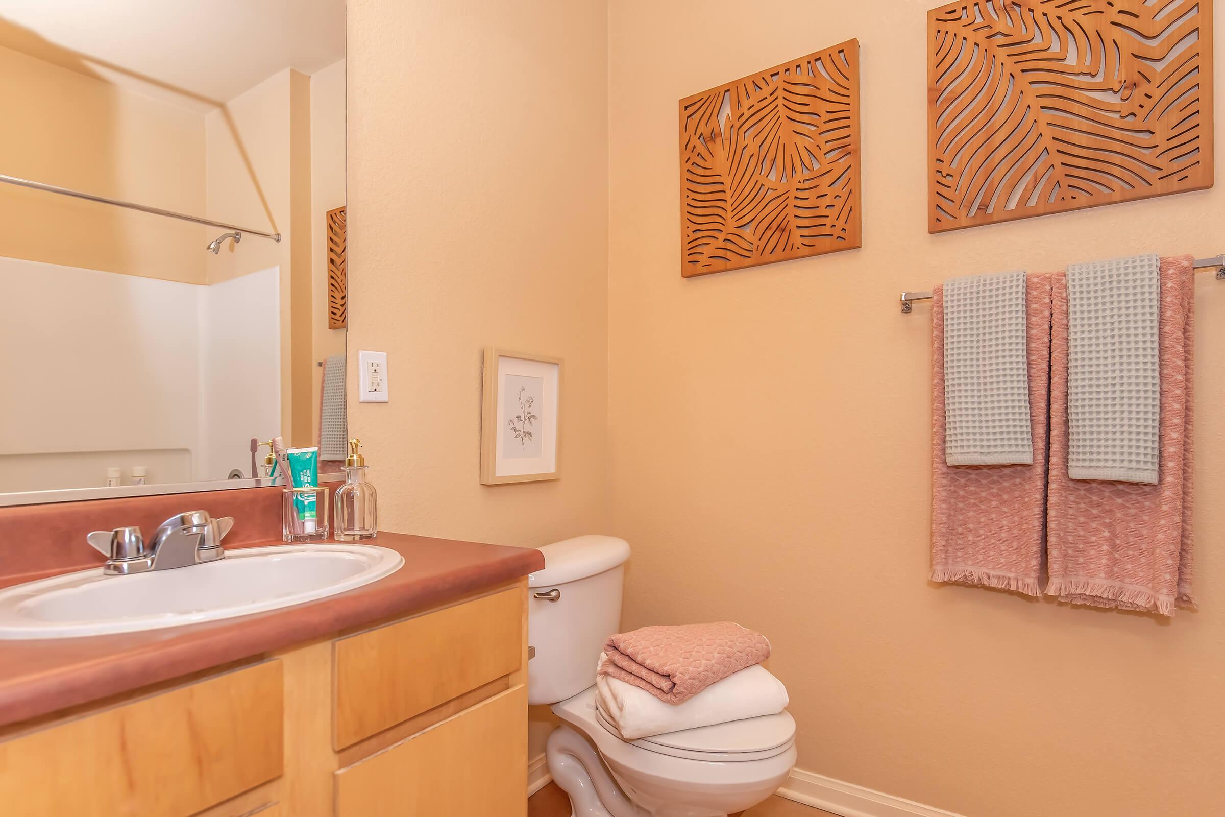 A modern bathroom featuring a sink with a wooden countertop, a white toilet, and neatly stacked towels. Two decorative wall hangings with a leaf pattern are above the sink. Soft pastel colors and natural light create a warm and inviting atmosphere.