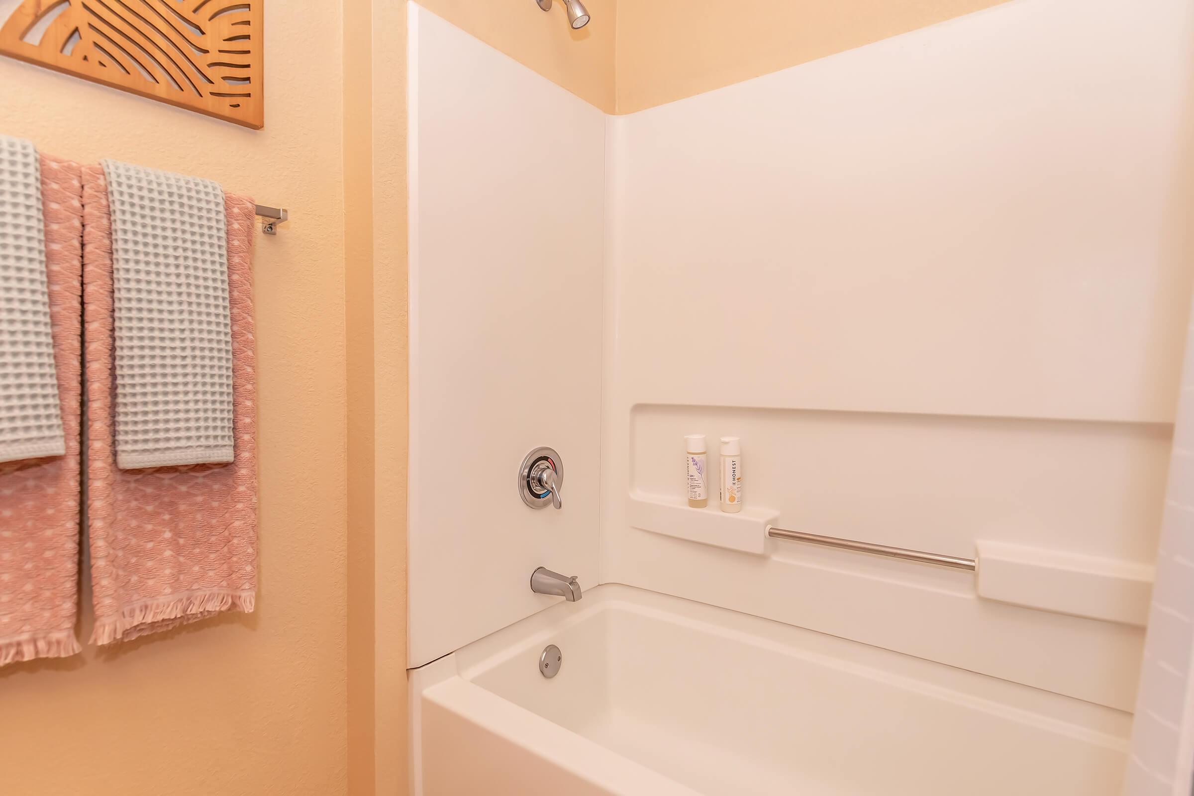 A clean bathroom with a light-colored wall, featuring a bathtub and a shower. On the wall, there are neatly hung towels in soft colors, and two bottles of toiletries are placed on a ledge next to the tub. The overall atmosphere is calming and well-organized.