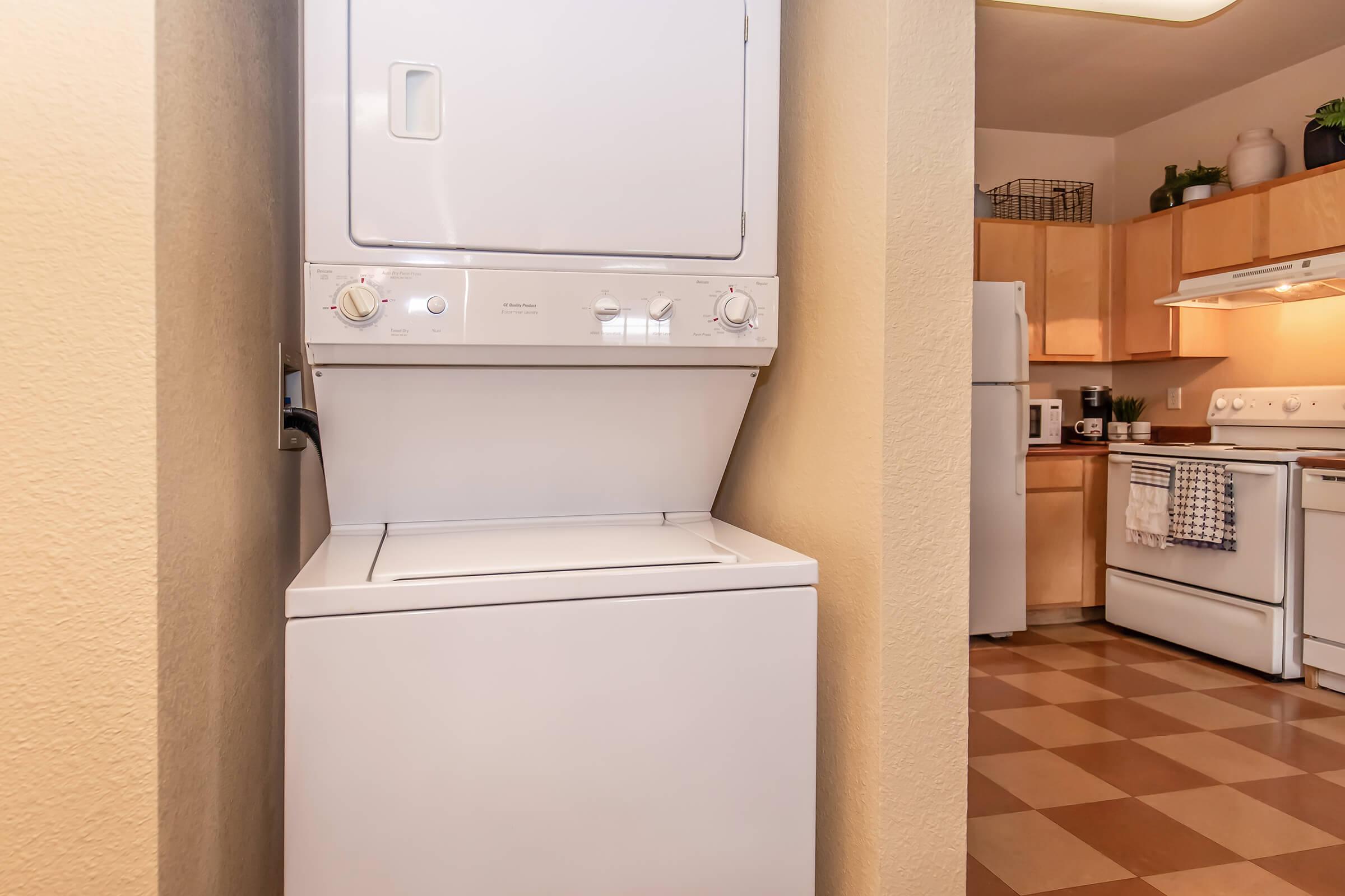 A compact laundry area featuring a stacked washer and dryer unit located between a wall and kitchen space. In the background, a well-equipped kitchen with wooden cabinets, a stove, and a refrigerator can be seen. The floor is tiled in a checkerboard pattern.