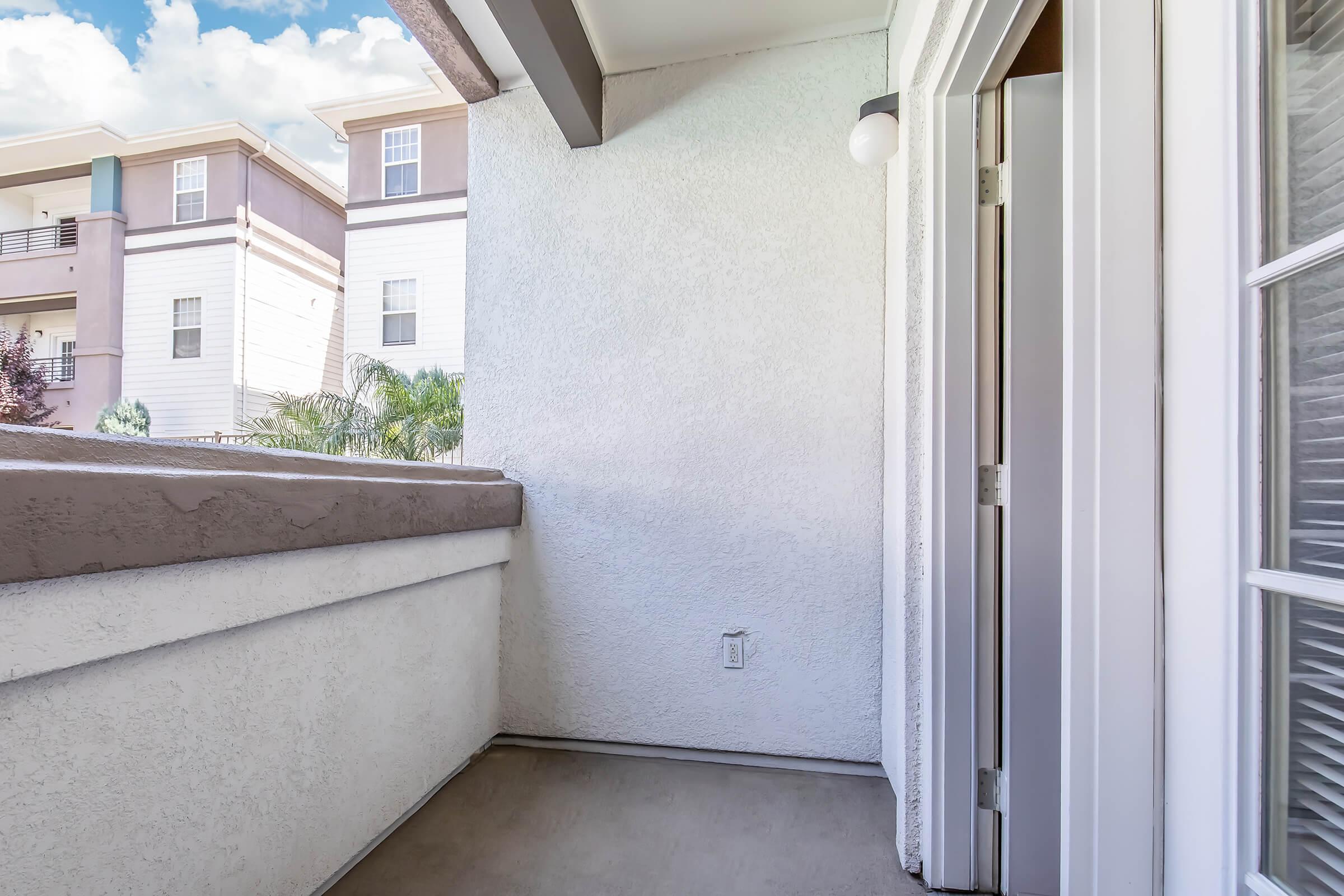 A small balcony with concrete flooring, featuring a white textured wall and a light fixture. The view includes the exterior of nearby buildings and palm trees in the background under a partly cloudy sky. The balcony has an open doorway leading to the interior space.