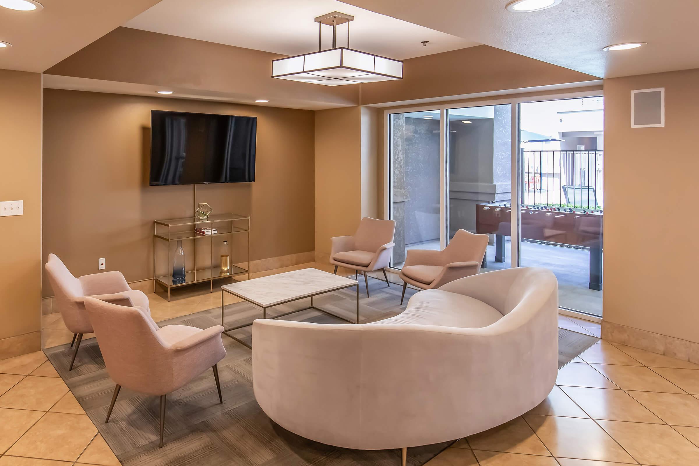 A modern living area featuring a curved white sofa and four light pink chairs surrounding a rectangular coffee table. A flat-screen TV is mounted on the wall, and large windows provide natural light, showcasing a view of an outdoor space. The walls are painted in a warm beige tone.