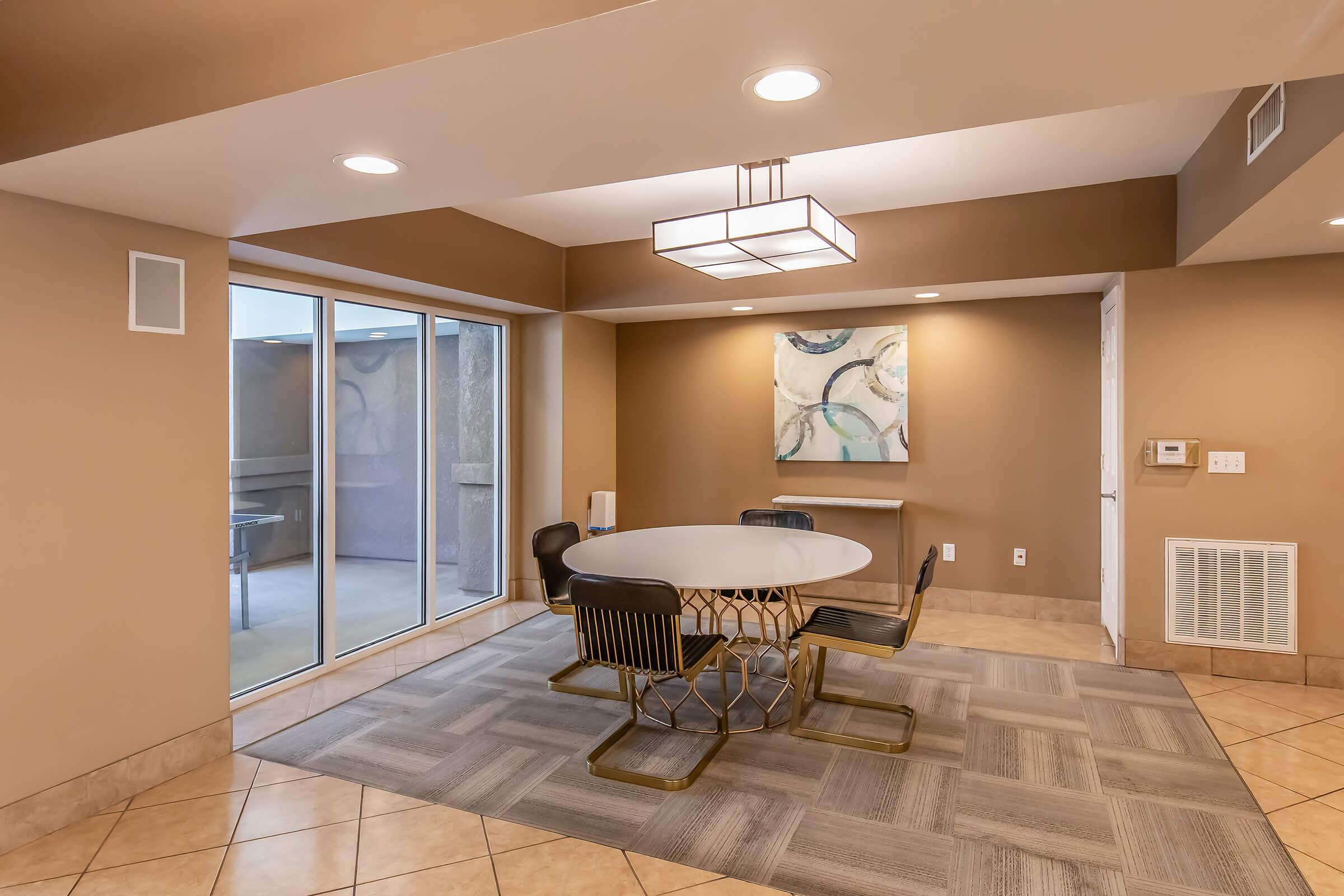 Modern dining area with a circular table surrounded by four black chairs. The walls are painted a warm beige color, and there's a large window providing natural light. A decorative pendant light hangs from the ceiling, and an abstract painting is mounted on the wall. The flooring features a light tile pattern.