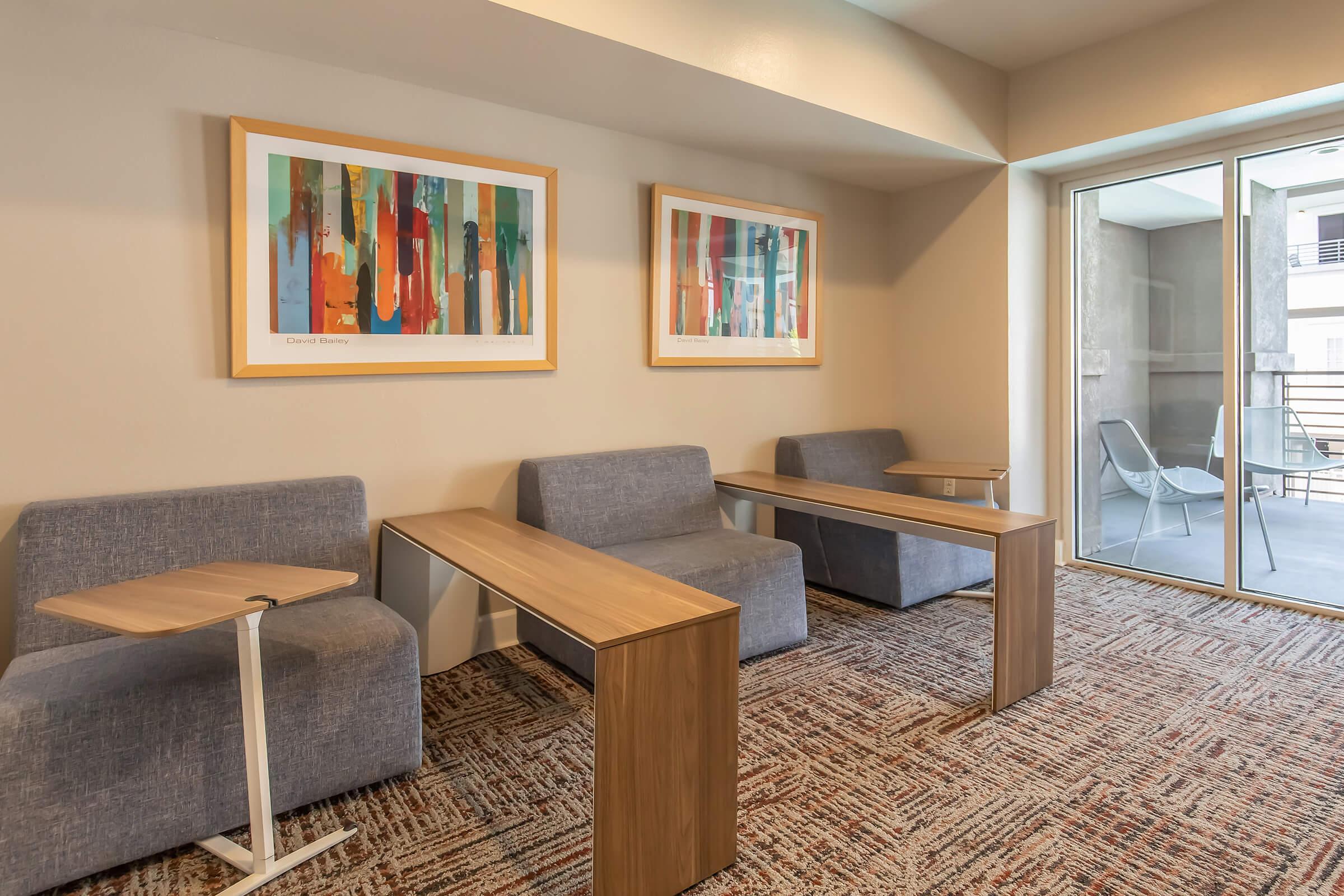 A modern lounge area featuring two gray sofas and minimalist wooden tables. Two framed artworks with vibrant abstract designs hang on the wall. A large window leads to an outdoor balcony area with additional seating. The flooring is covered with a patterned carpet, creating a cozy atmosphere.
