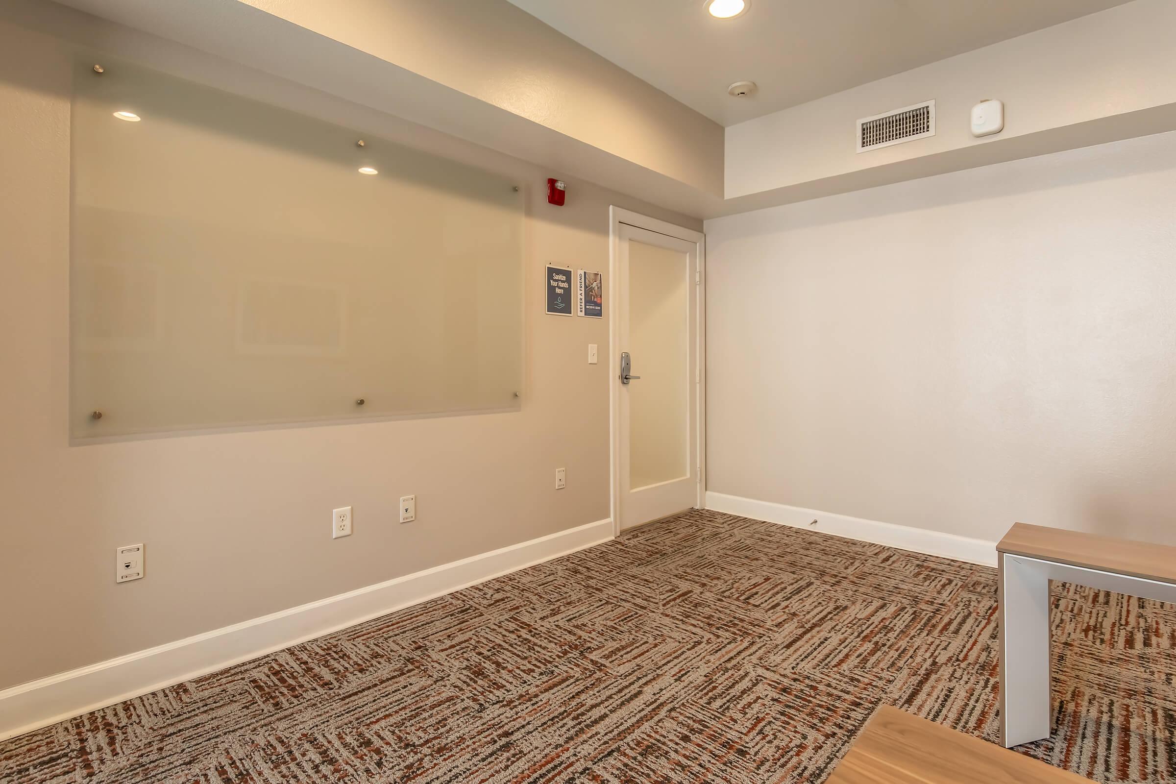 A modern, empty room with light gray walls and carpet. On one wall, there's a frosted glass board with no writing. A door leads to another space, and there's a small wooden table in the room. The overall atmosphere is minimalistic and clean, with soft lighting.