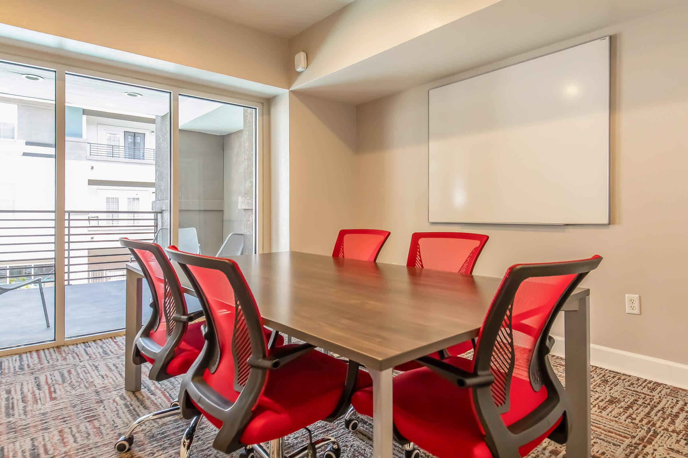 A modern conference room featuring a large rectangular table surrounded by six red office chairs. A whiteboard is mounted on the wall, and large windows provide natural light, with a view of a balcony outside. The floor is covered in a patterned carpet, creating a professional yet inviting atmosphere.