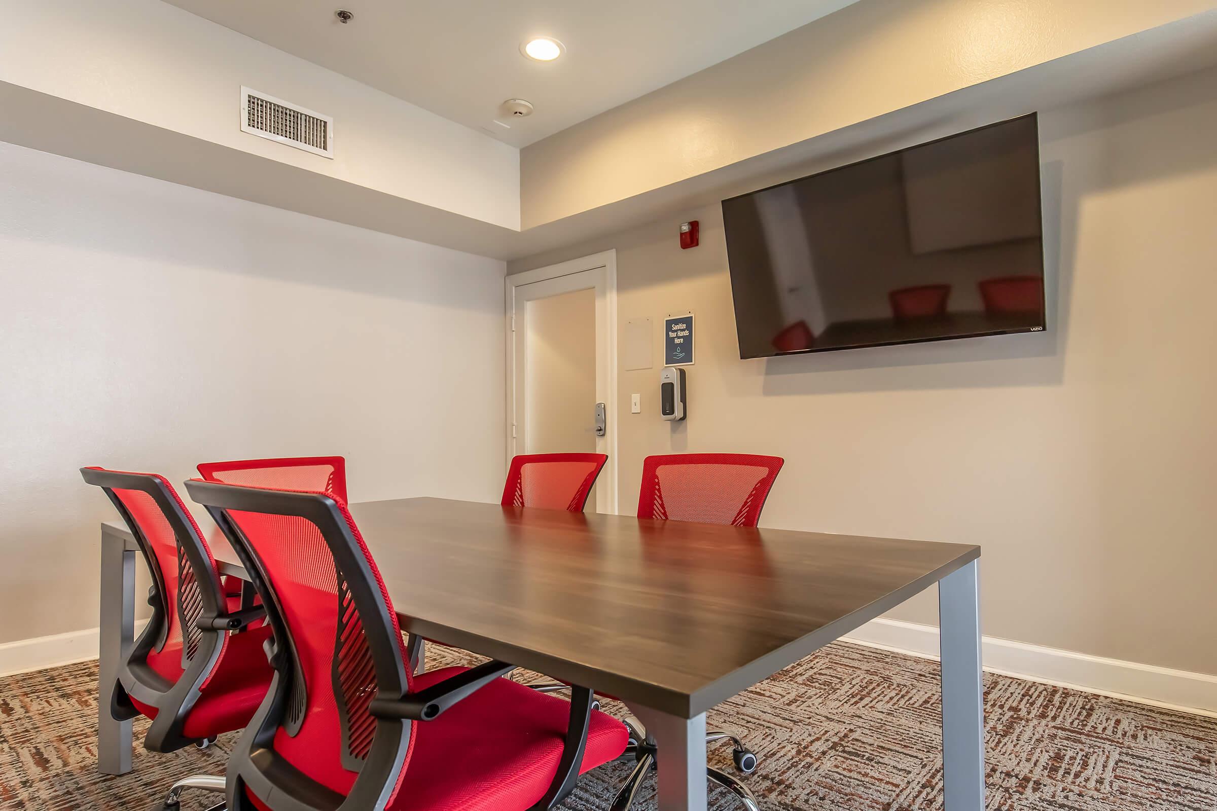 A modern conference room featuring a large wooden table surrounded by five red mesh chairs. A flat-screen television is mounted on the wall, and there is a door leading to another space. The room has neutral-colored walls and carpet, providing a professional atmosphere suitable for meetings.