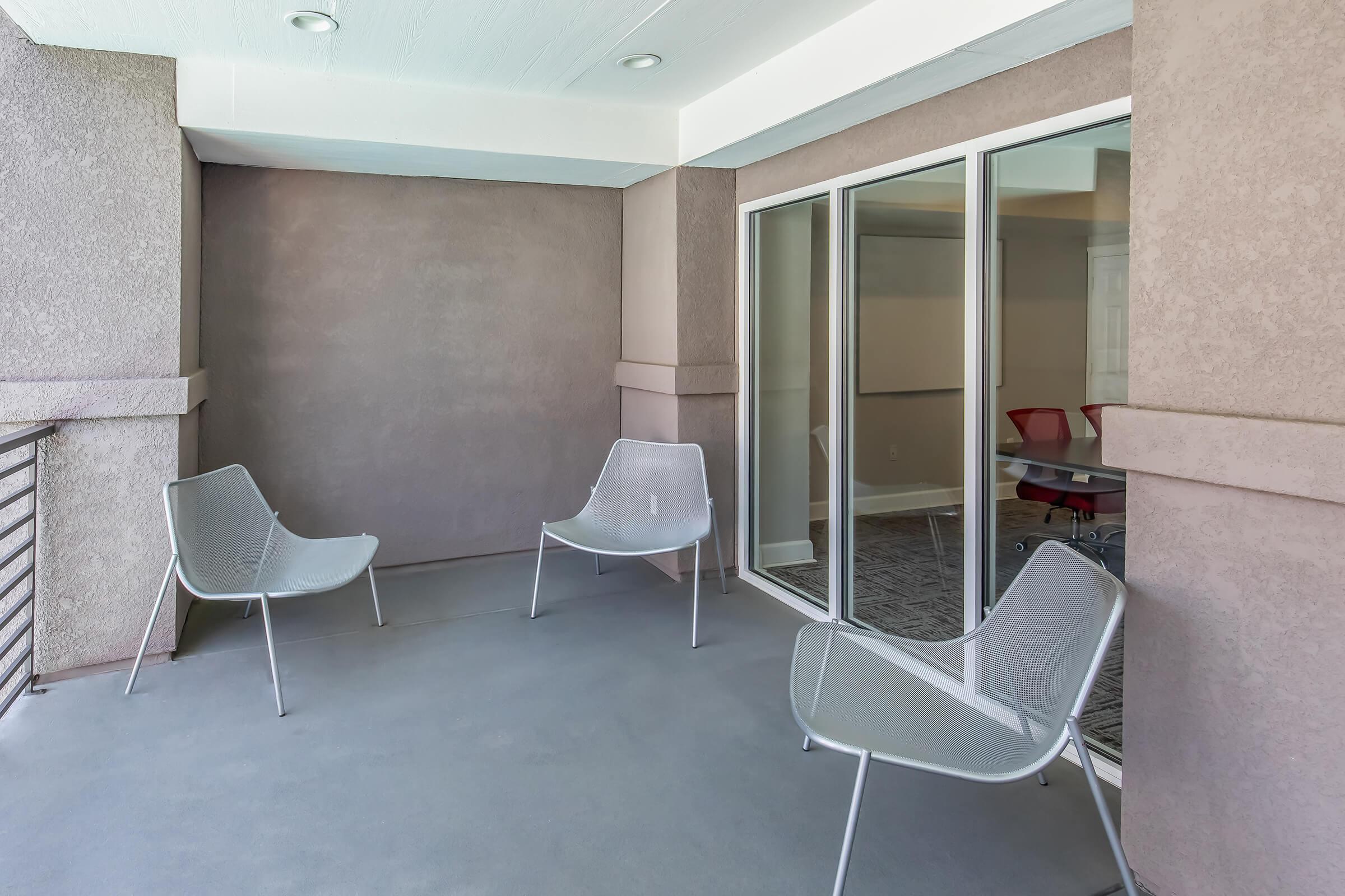 A spacious balcony featuring four modern, lightweight chairs arranged around a simple, minimalist outdoor setting. The backdrop includes large glass windows reflecting an indoor meeting space, with neutral-toned walls and a smooth concrete floor, creating an inviting atmosphere for relaxation or conversation.