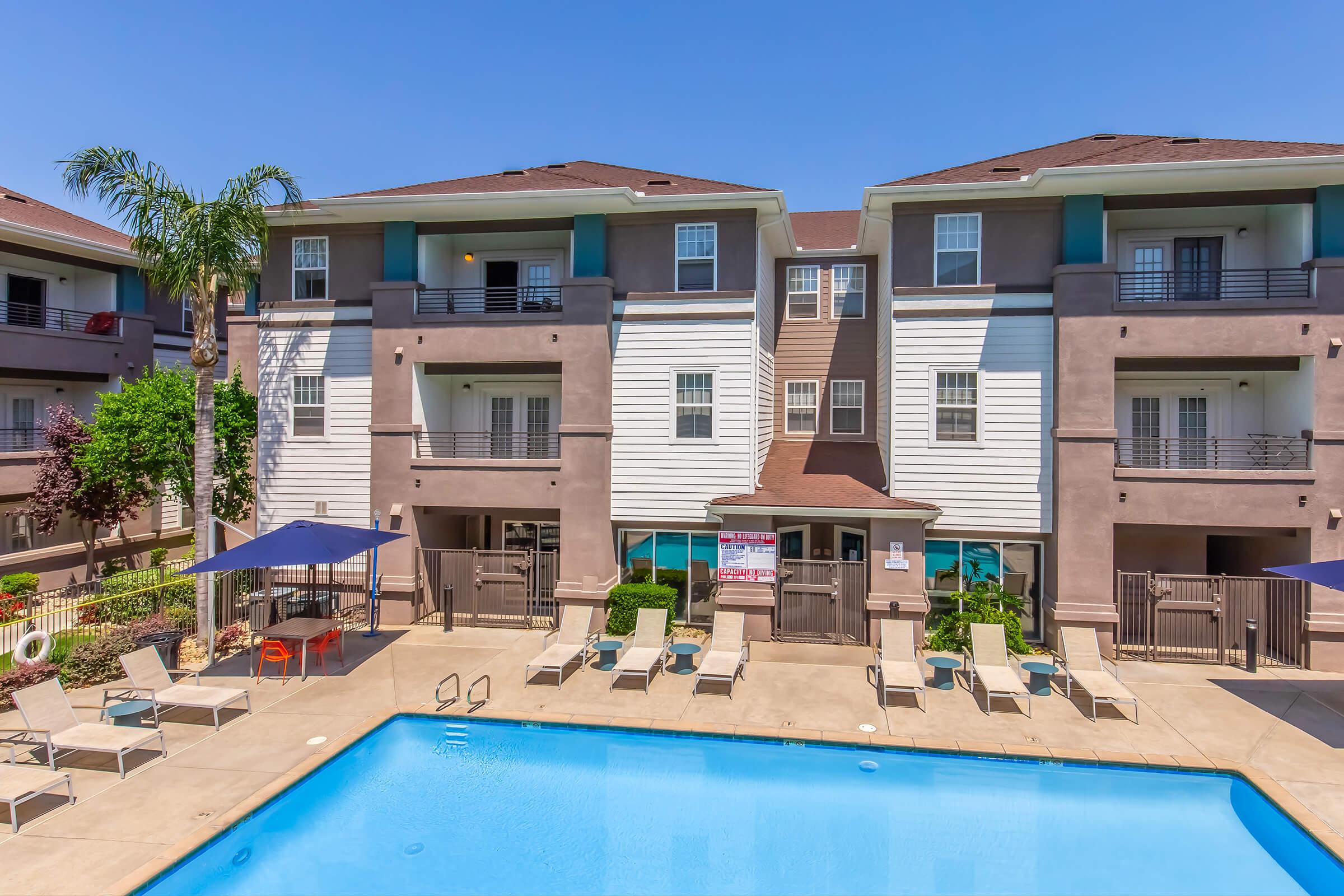 A swimming pool surrounded by lounge chairs, with an apartment building in the background. The building features multiple levels, balconies, and landscaped greenery. Clear blue sky above enhances the sunny atmosphere of the pool area.