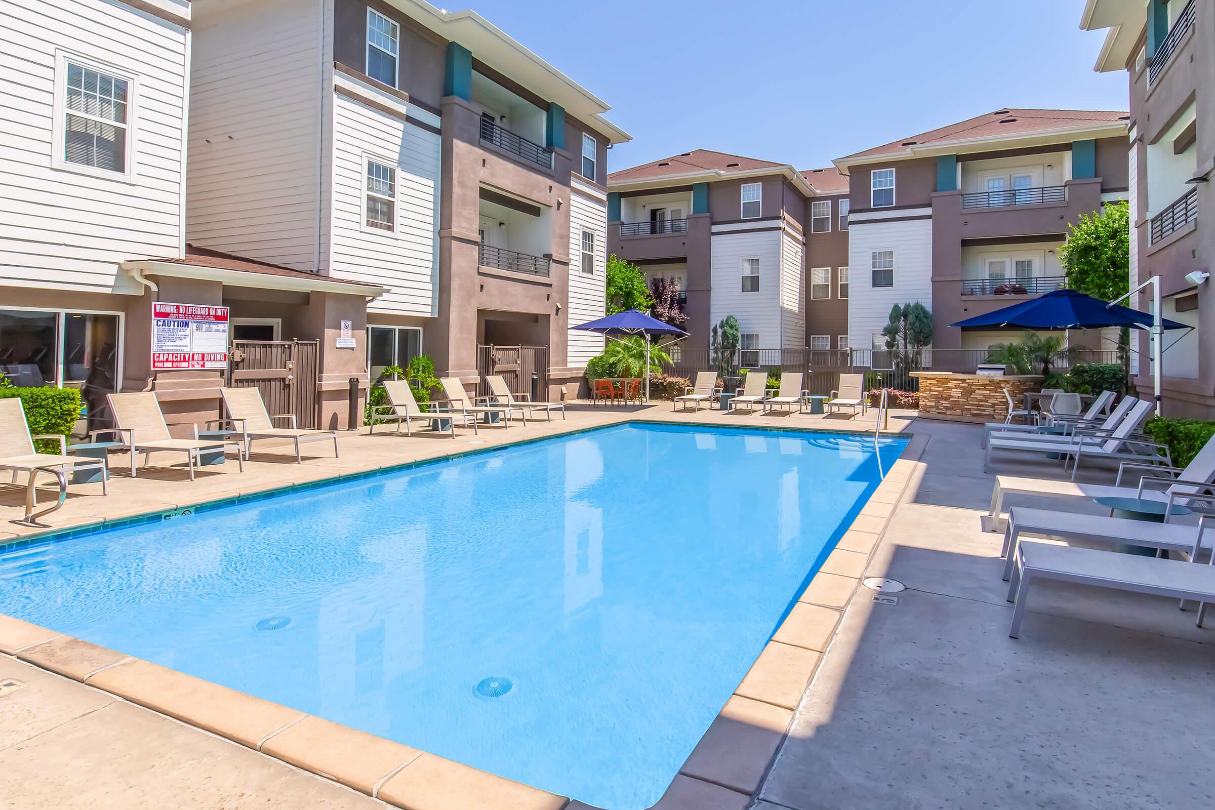 A clean and inviting outdoor swimming pool surrounded by lounge chairs and umbrellas, set within a modern apartment complex. The buildings are two to three stories high, featuring a mix of light and dark siding. Lush greenery is visible around the pool area under a clear blue sky.