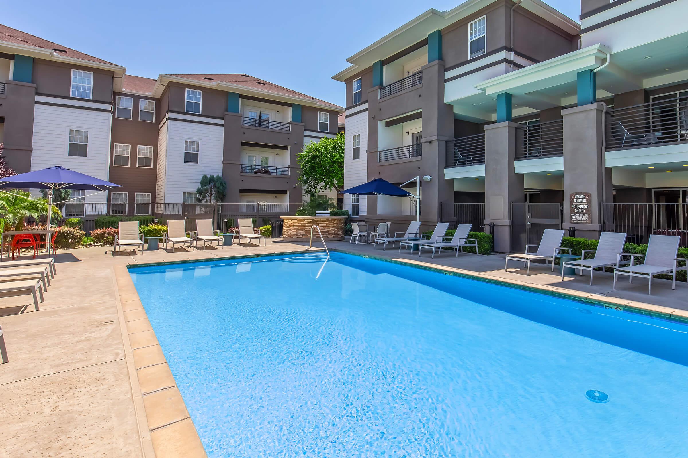 A bright, inviting swimming pool surrounded by lounge chairs, with modern apartment buildings in the background. The pool area features umbrellas for shade and landscaped greenery, creating a relaxing outdoor environment for residents and guests.