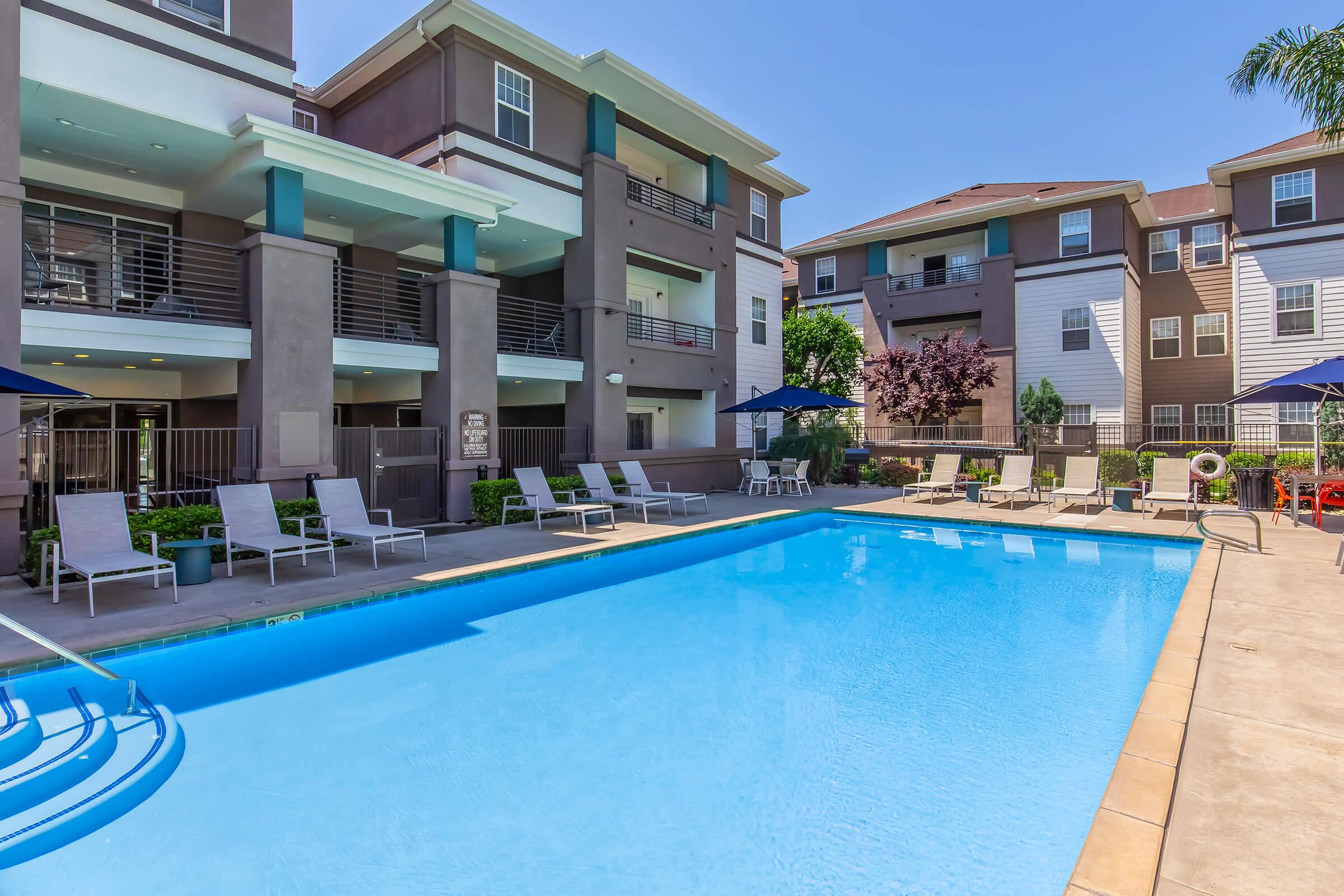 A clear blue swimming pool surrounded by lounge chairs and umbrellas, situated in an outdoor area of an apartment complex. The buildings in the background have a modern design with balconies and greenery, creating a relaxing atmosphere. Bright sunny day with a clear blue sky.