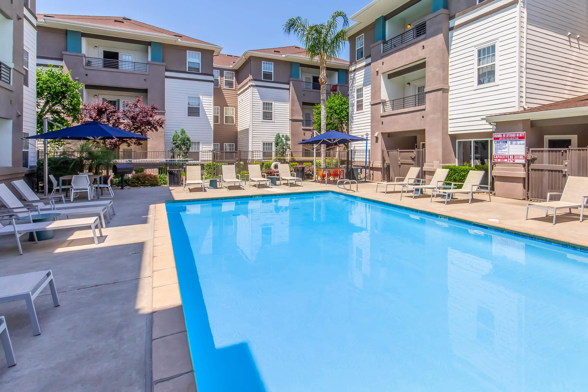 A clear blue swimming pool surrounded by sun loungers and umbrellas, set within a courtyard of modern apartment buildings. Lush greenery and trees are visible, creating a relaxing atmosphere. The sunny day highlights the pool's inviting water, making it an appealing spot for relaxation.
