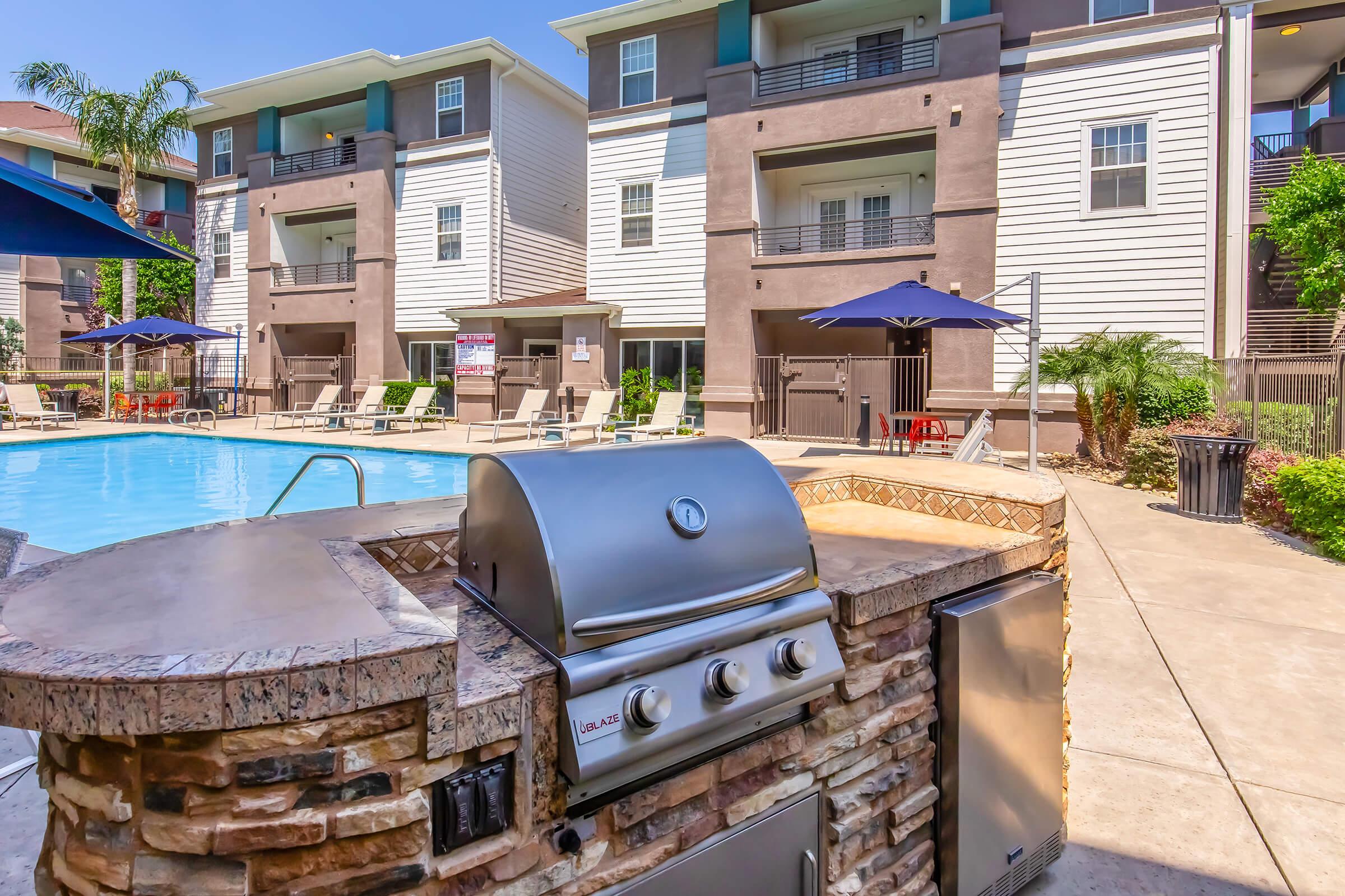 A modern apartment complex features a swimming pool surrounded by lounge chairs and umbrellas. In the foreground, a built-in outdoor grill is set on a stone countertop, providing a space for barbecuing. Lush greenery and architectural details of the buildings enhance the inviting atmosphere.