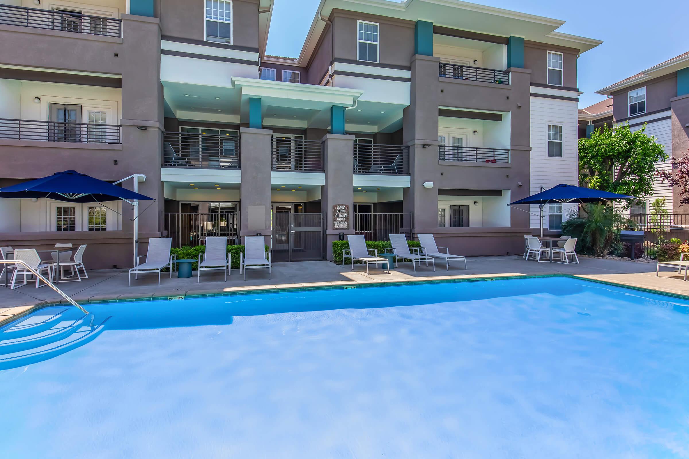 View of a modern apartment complex featuring a swimming pool in the foreground. The pool is surrounded by lounge chairs and umbrellas. The building has multiple stories with balconies and large windows, set in a well-maintained outdoor area with greenery.